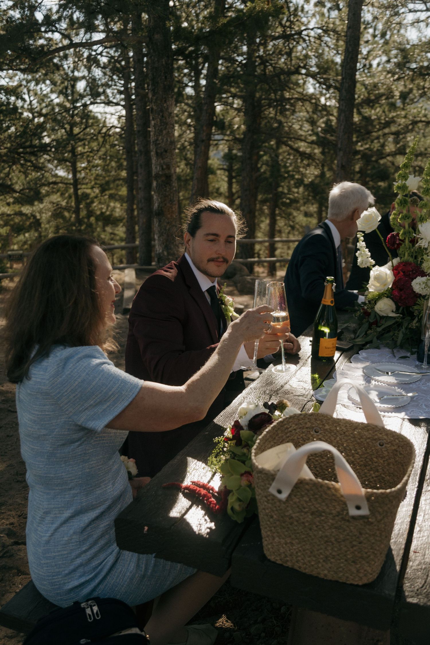 Couple toasting champagne glasses at outdoor woodland wedding reception table with floral arrangements.