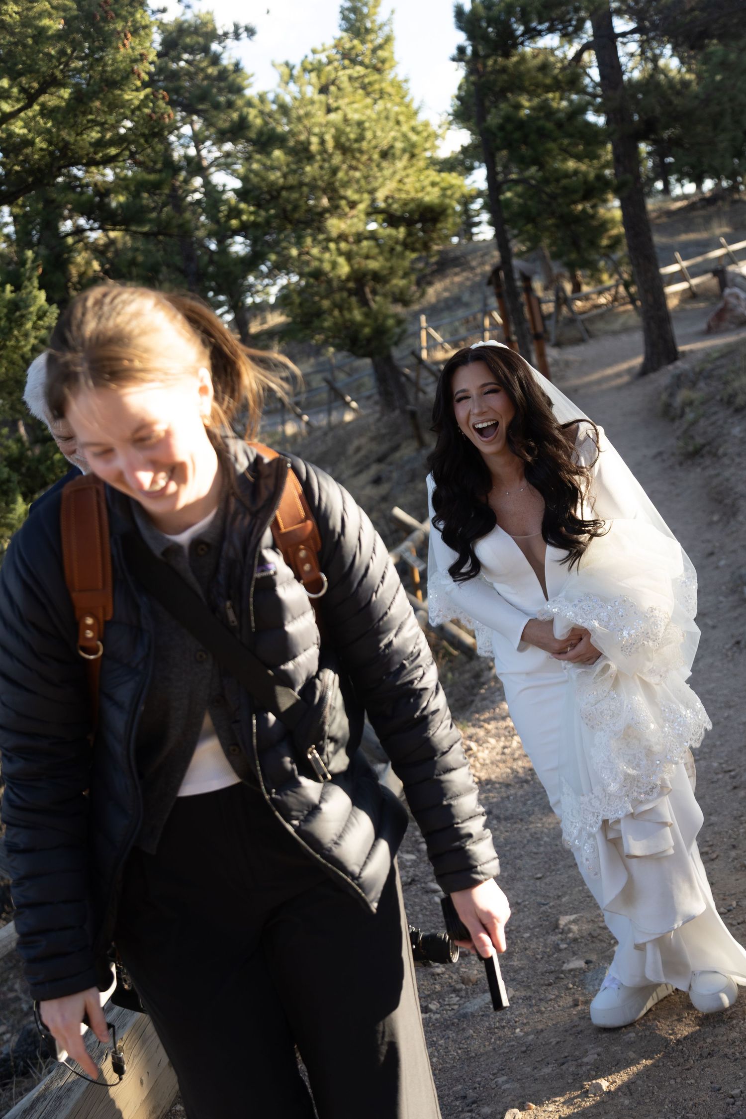 Bride in white gown laughs with a guest on a forested mountain trail during wedding celebration.