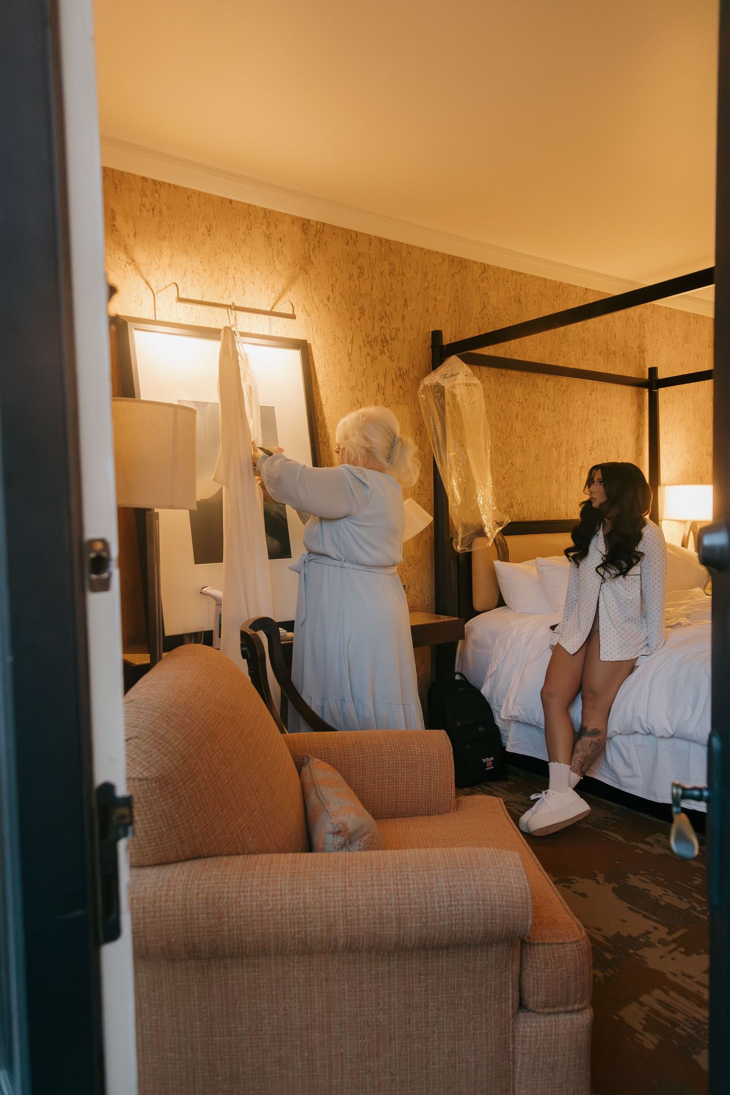 Mother helps bride with wedding dress in warm hotel room with four-poster bed before ceremony.