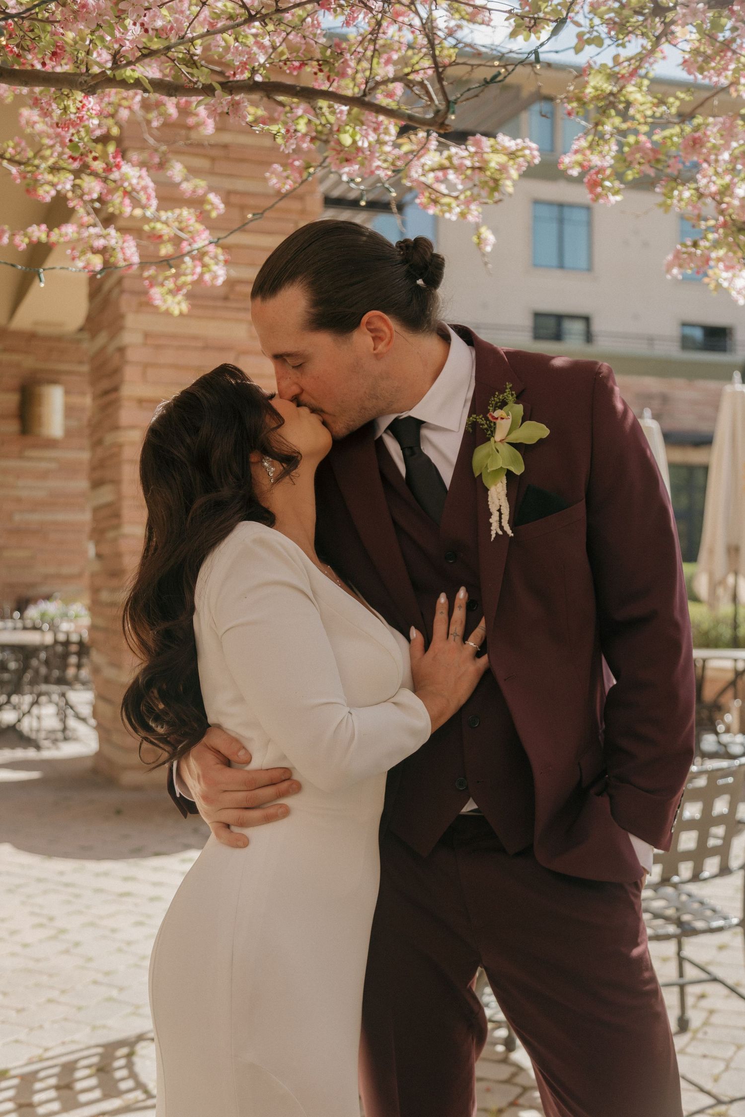 Bride and groom kissing under blooming pink cherry blossom tree, groom in burgundy suit with boutonniere.