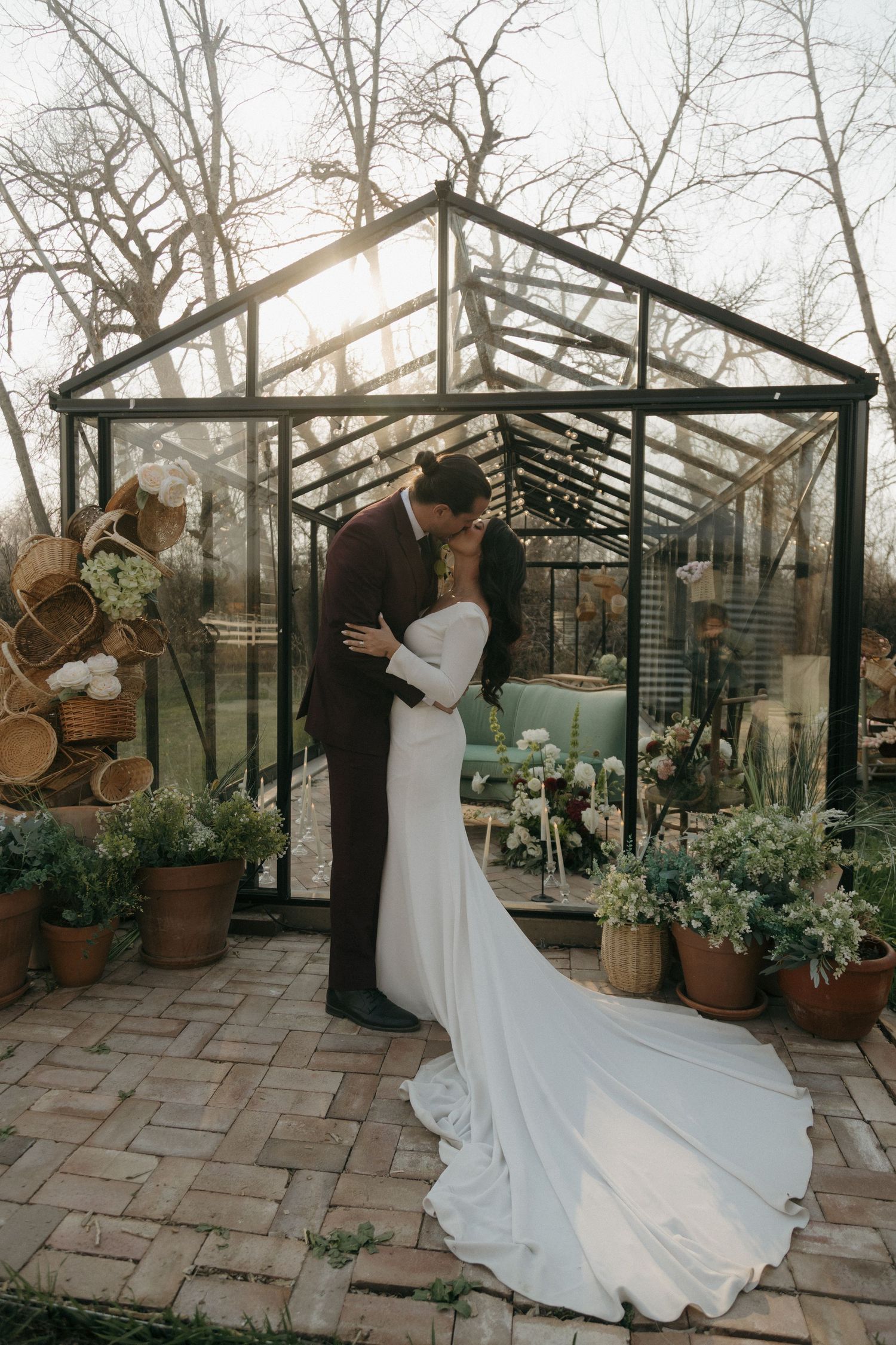 Bride and groom share a kiss in front of a glass greenhouse surrounded by potted plants at golden hour.