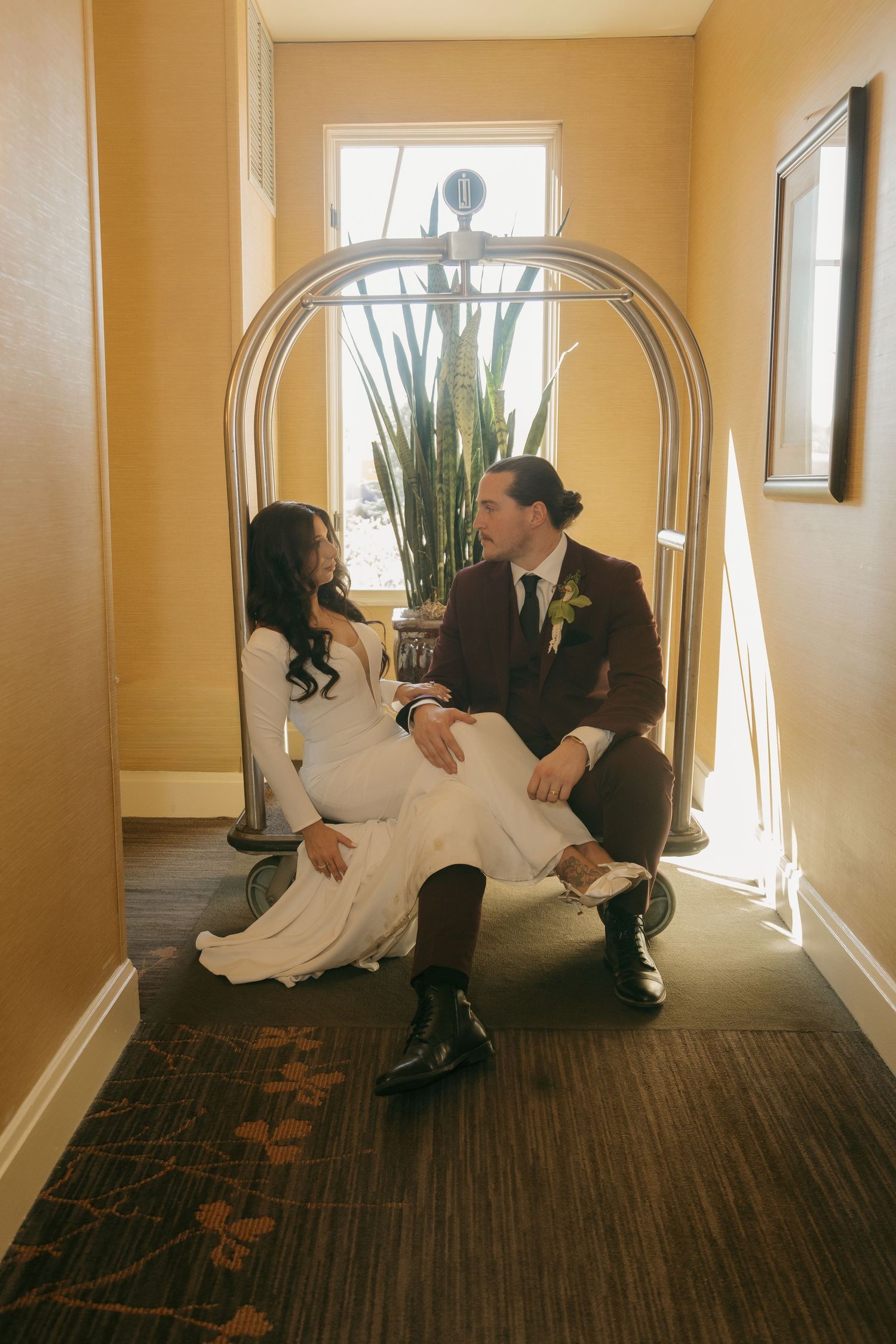 Bride and groom sitting together on a hotel luggage cart in a warmly lit corridor.