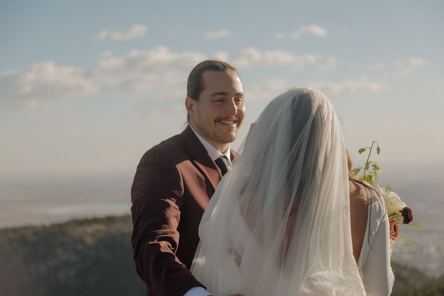 A smiling groom in a burgundy suit looks back at his veiled bride on a mountaintop.