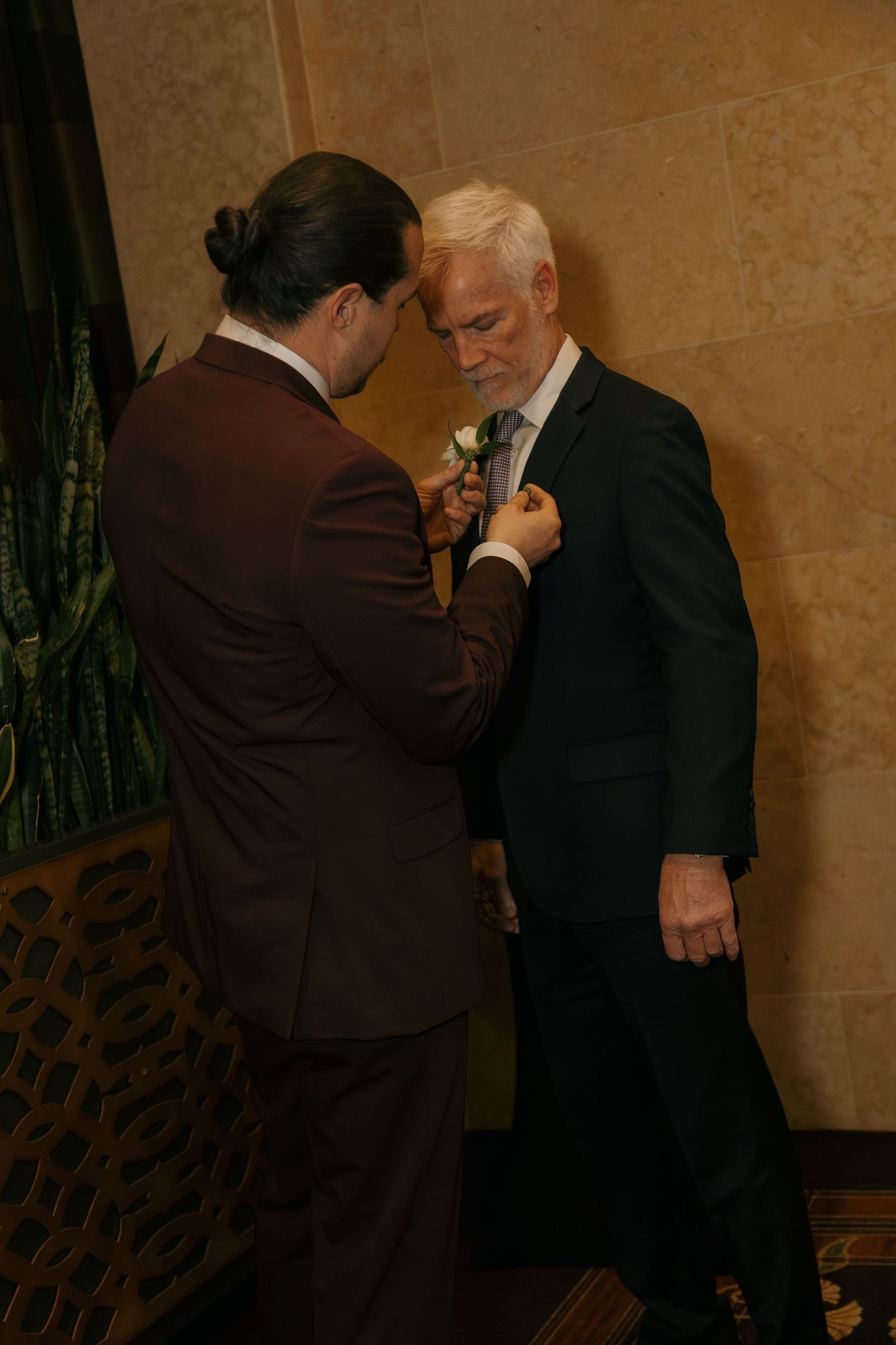 Man in burgundy suit pins boutonniere on older gentleman in dark suit at wedding.