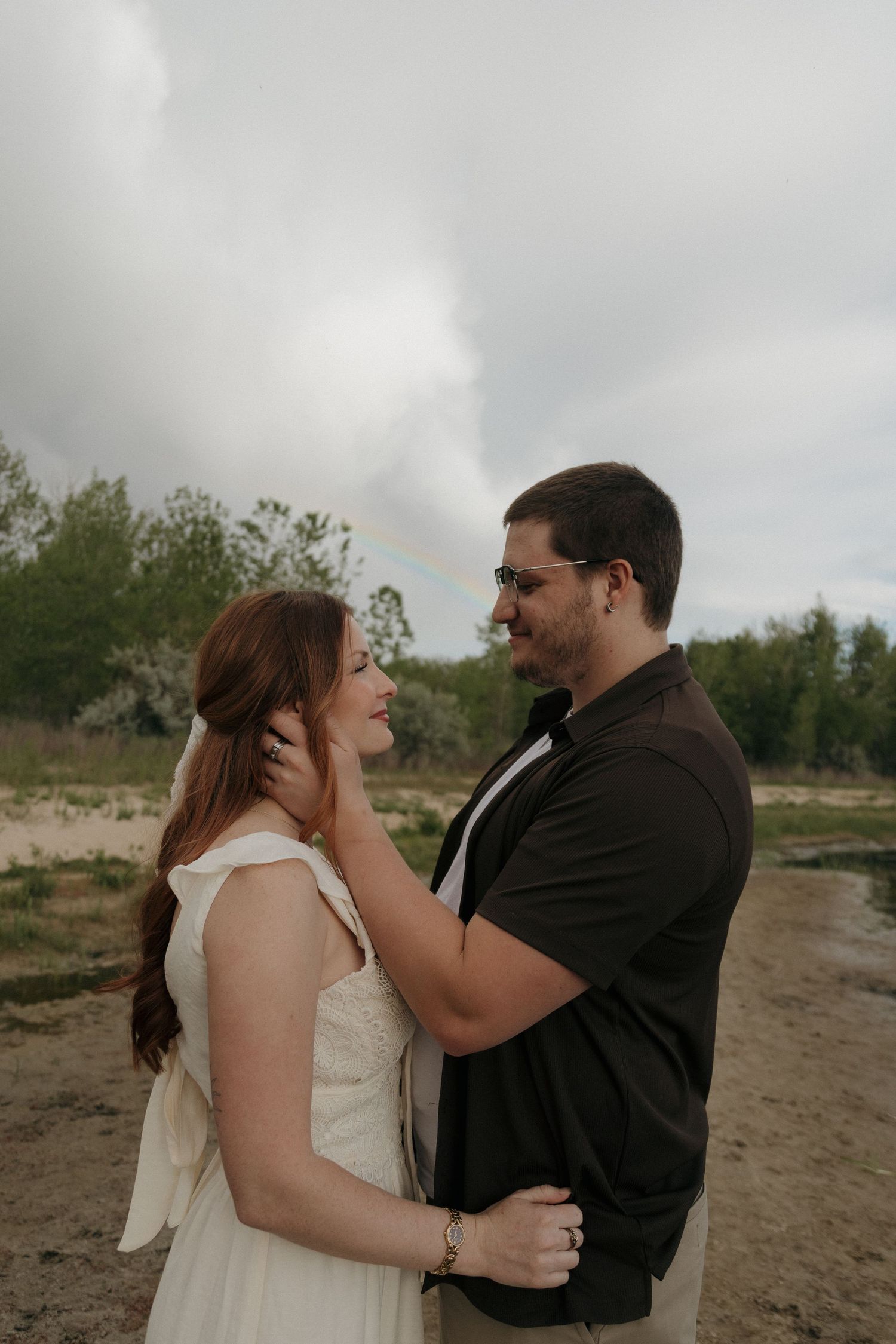 Romantic couple sharing a tender moment outdoors, man gently touching womans face in a white dress with stormy sky backdrop.