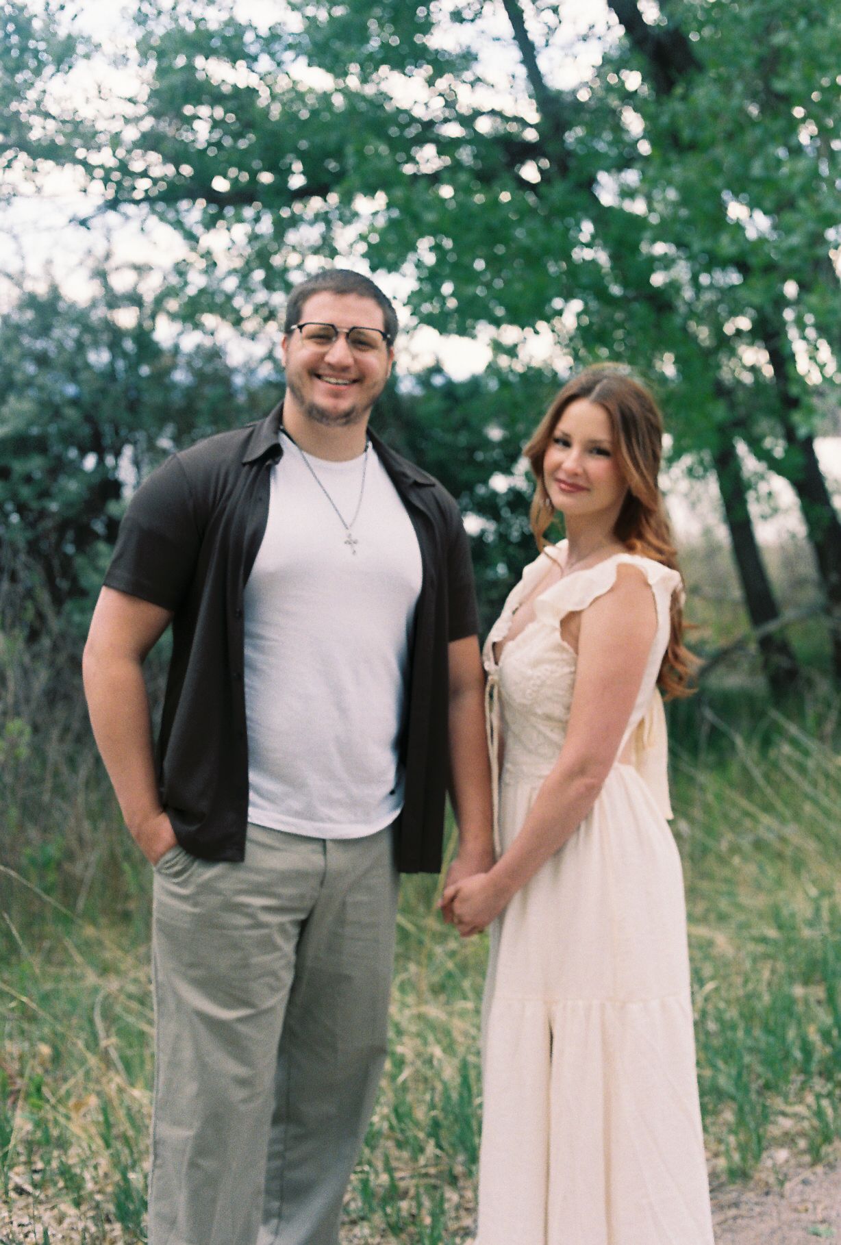 Smiling couple posing together in a wooded outdoor setting, man in black shirt and woman in white dress.