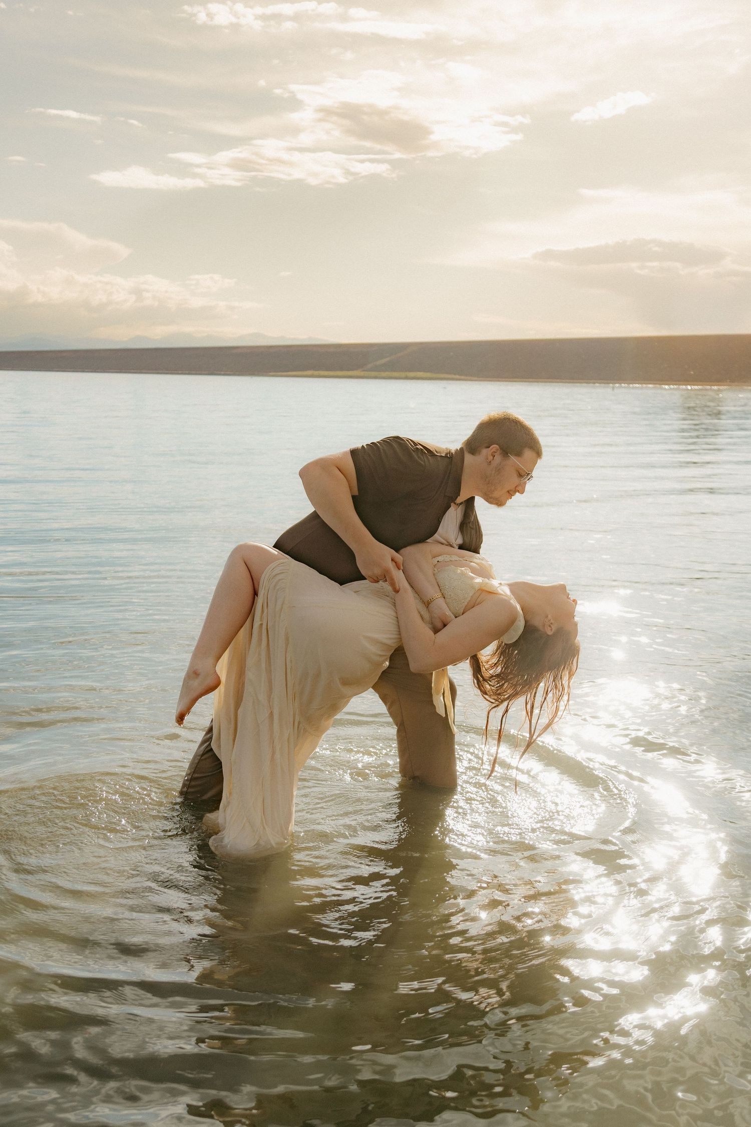 Man dipping woman in white dress dramatically over lake water during golden sunset.