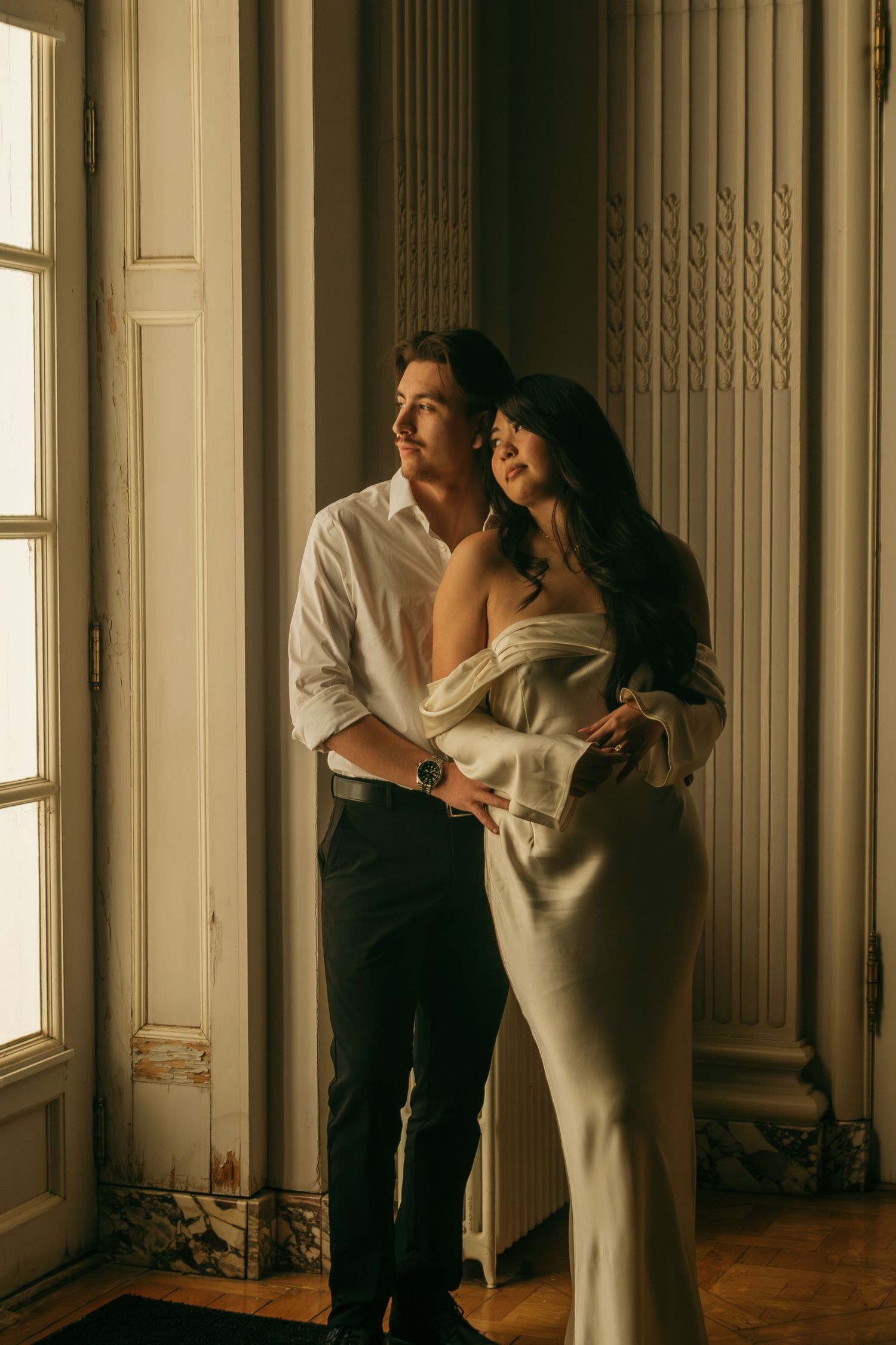 Elegant couple in formal attire embracing by a sunlit window in a grand interior setting.