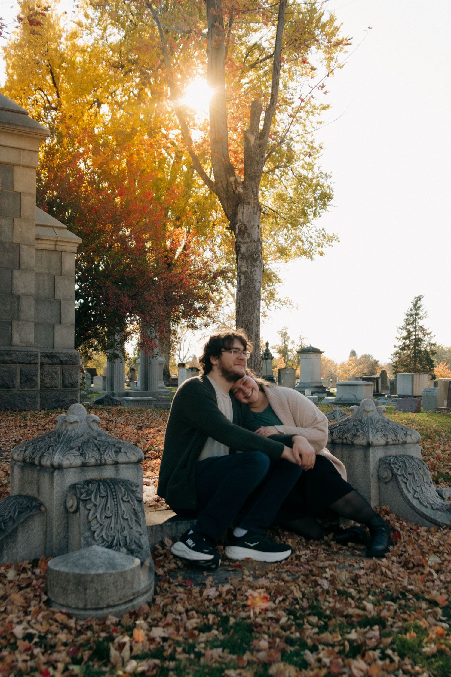 Couple sitting together among autumn leaves in a sunlit cemetery, surrounded by stone monuments and golden fall foliage.