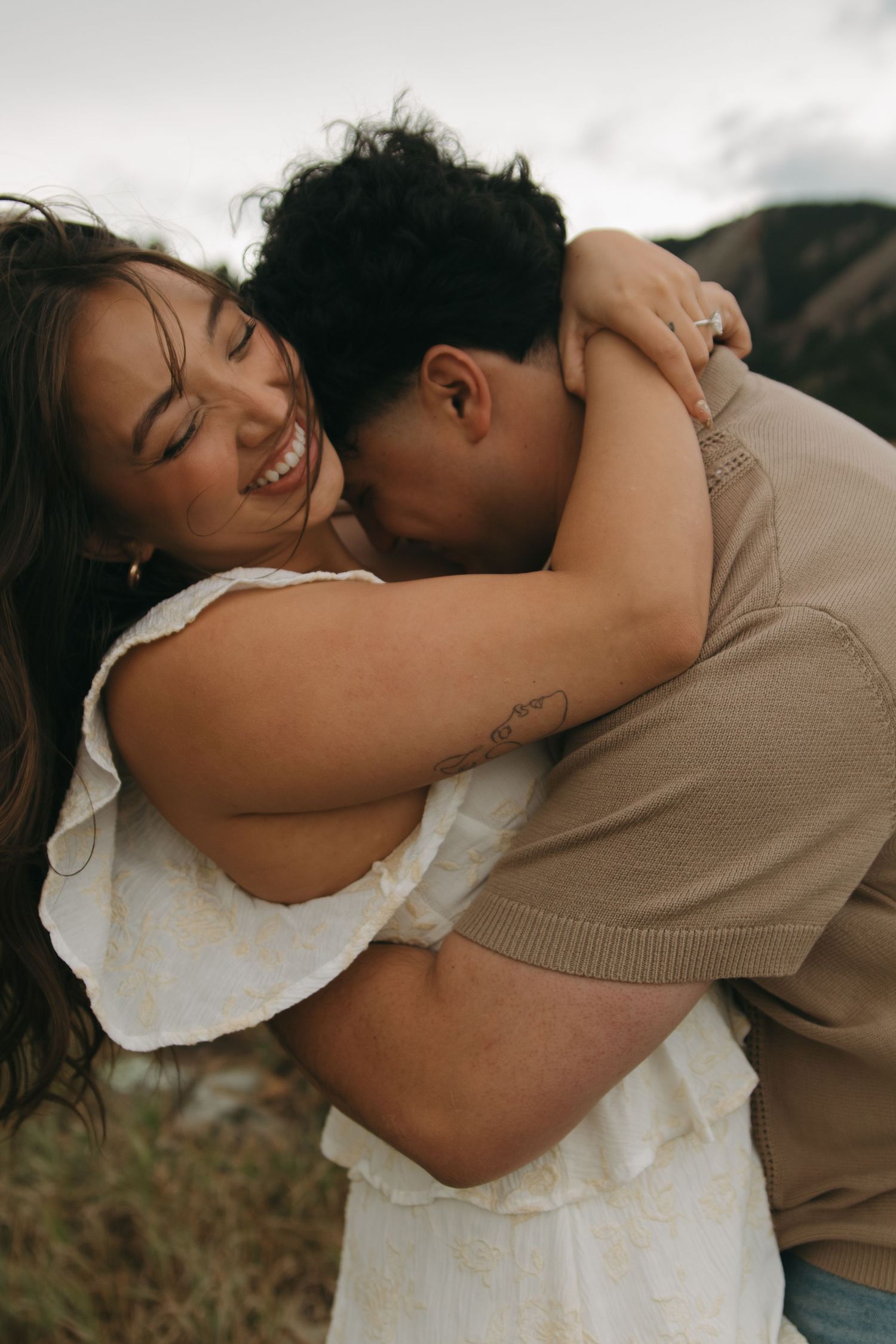 A couple shares a tender embrace outdoors, with the man kissing the smiling woman's cheek in a romantic moment.