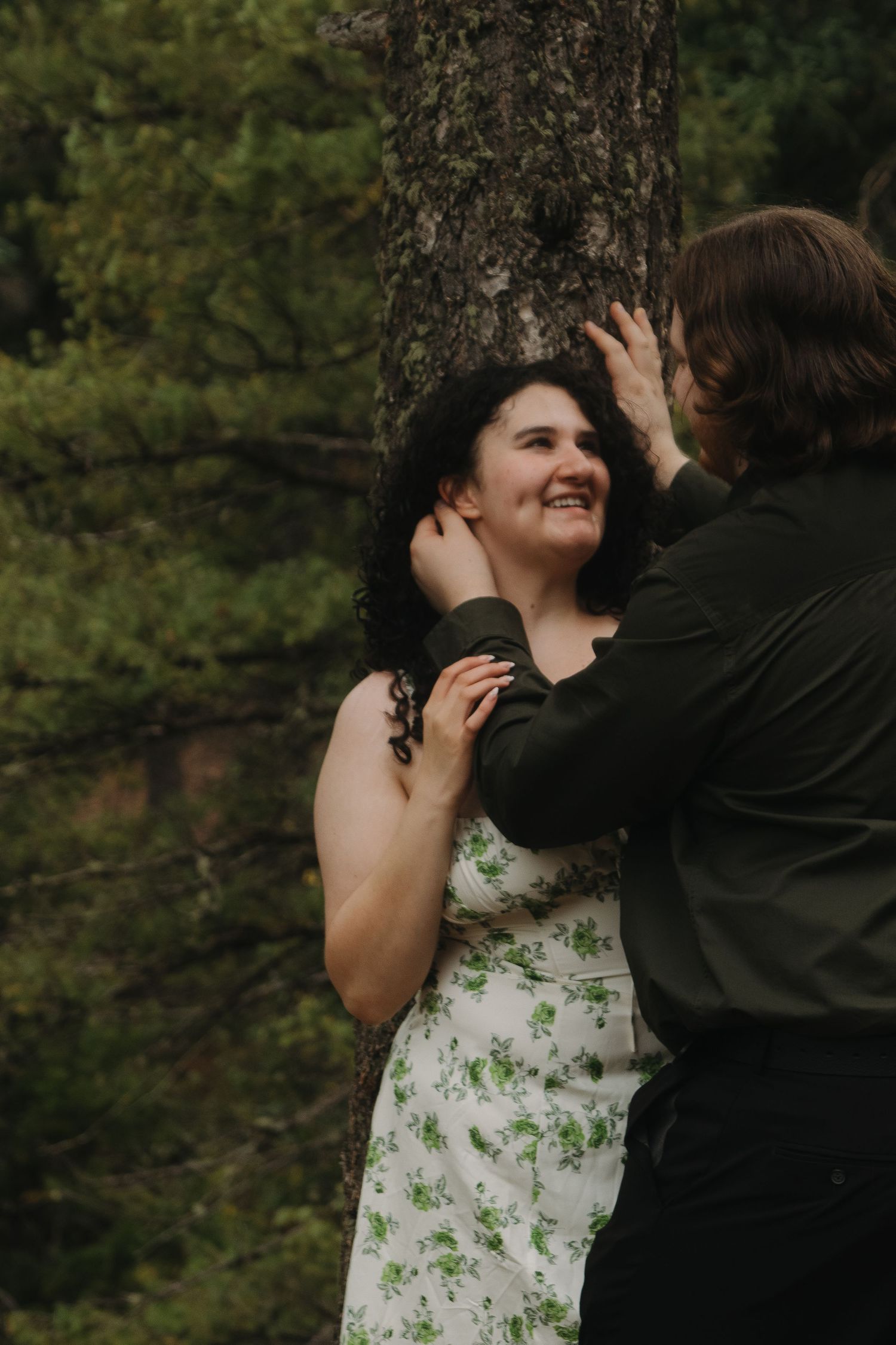 A romantic moment captured between a couple against a pine tree in a forest setting.