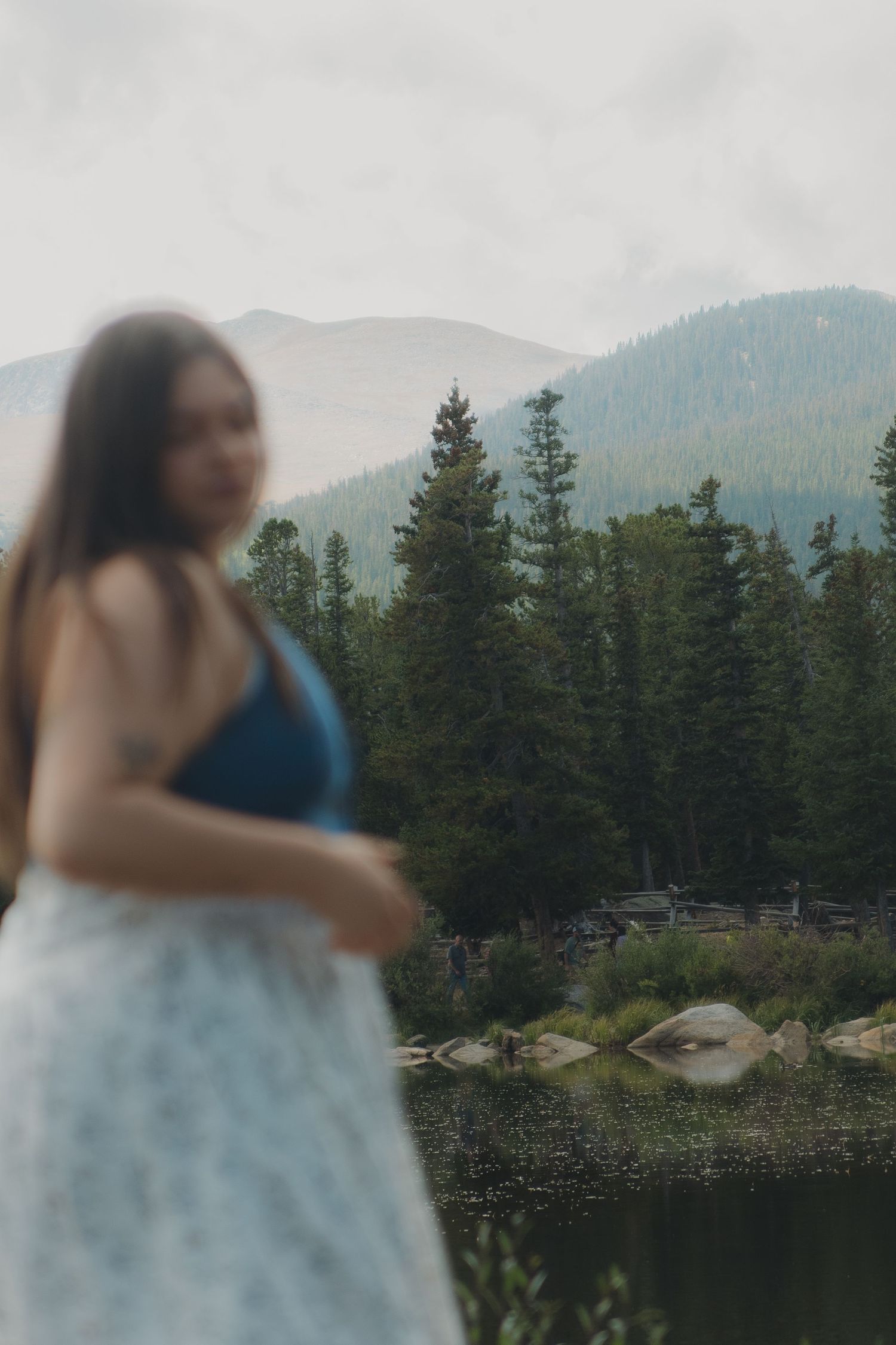 A scenic mountain lake surrounded by evergreen trees with a blurred figure in a blue and white dress in the foreground.