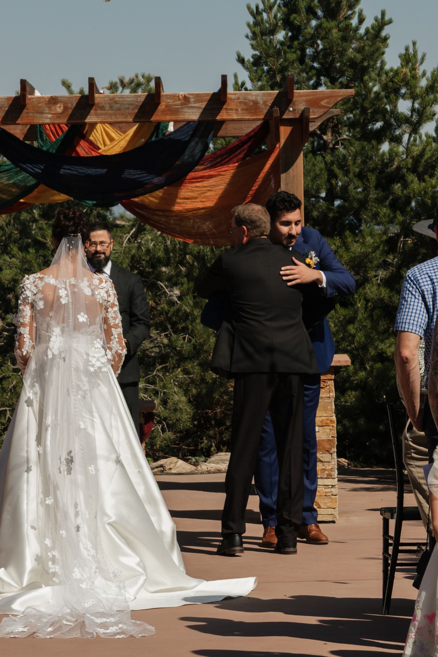 A wedding ceremony taking place under a wooden pergola with a flowing white wedding dress visible.