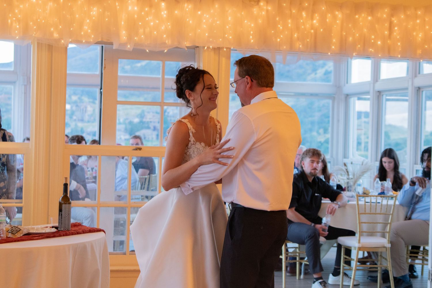 A wedding reception room glowing with warm string lights and large windows.