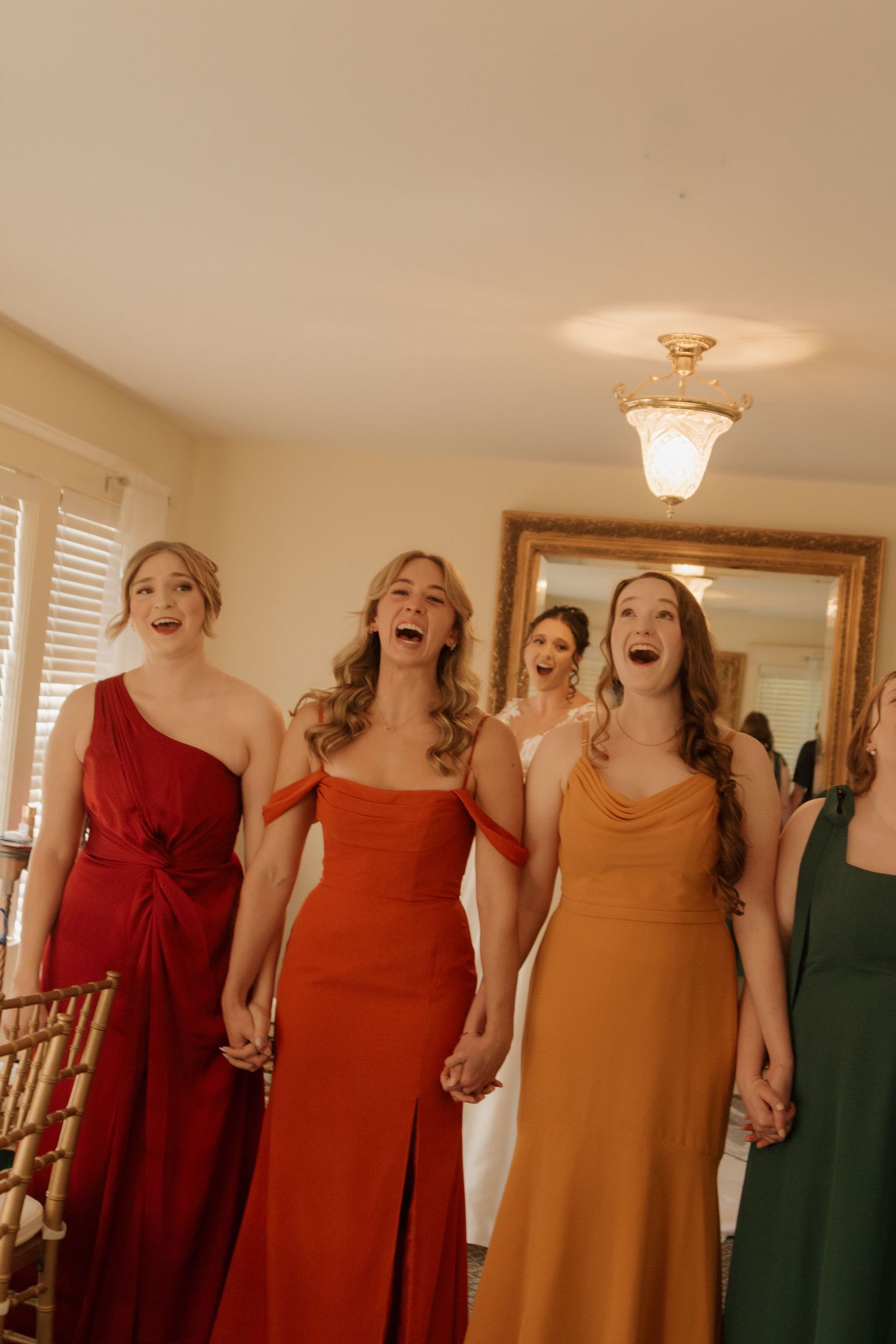 Bridesmaids in red and orange dresses laugh joyfully together during a wedding celebration.