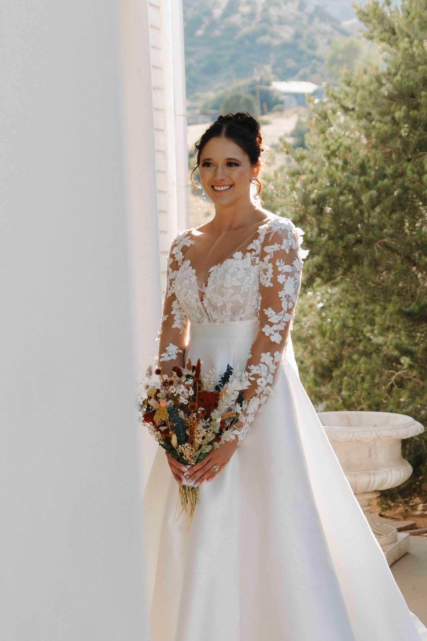 Elegant wedding dress with lace sleeves and bouquet against mountain backdrop.