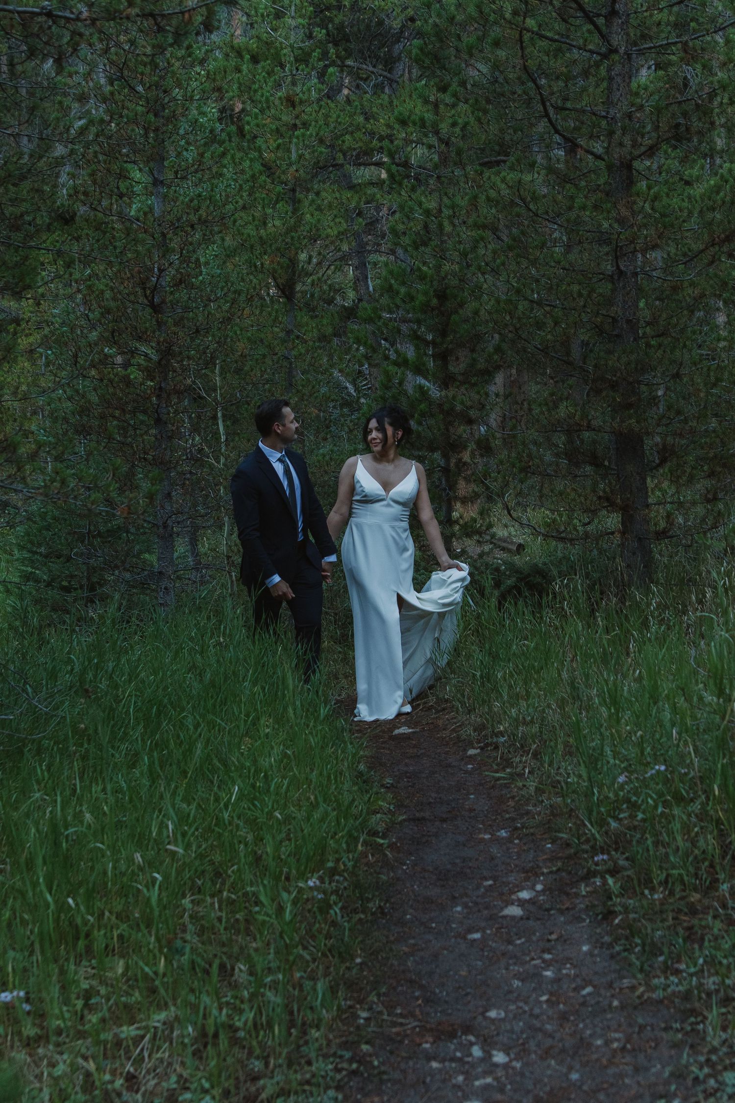 A couple in wedding attire walking along a forest trail surrounded by lush greenery.