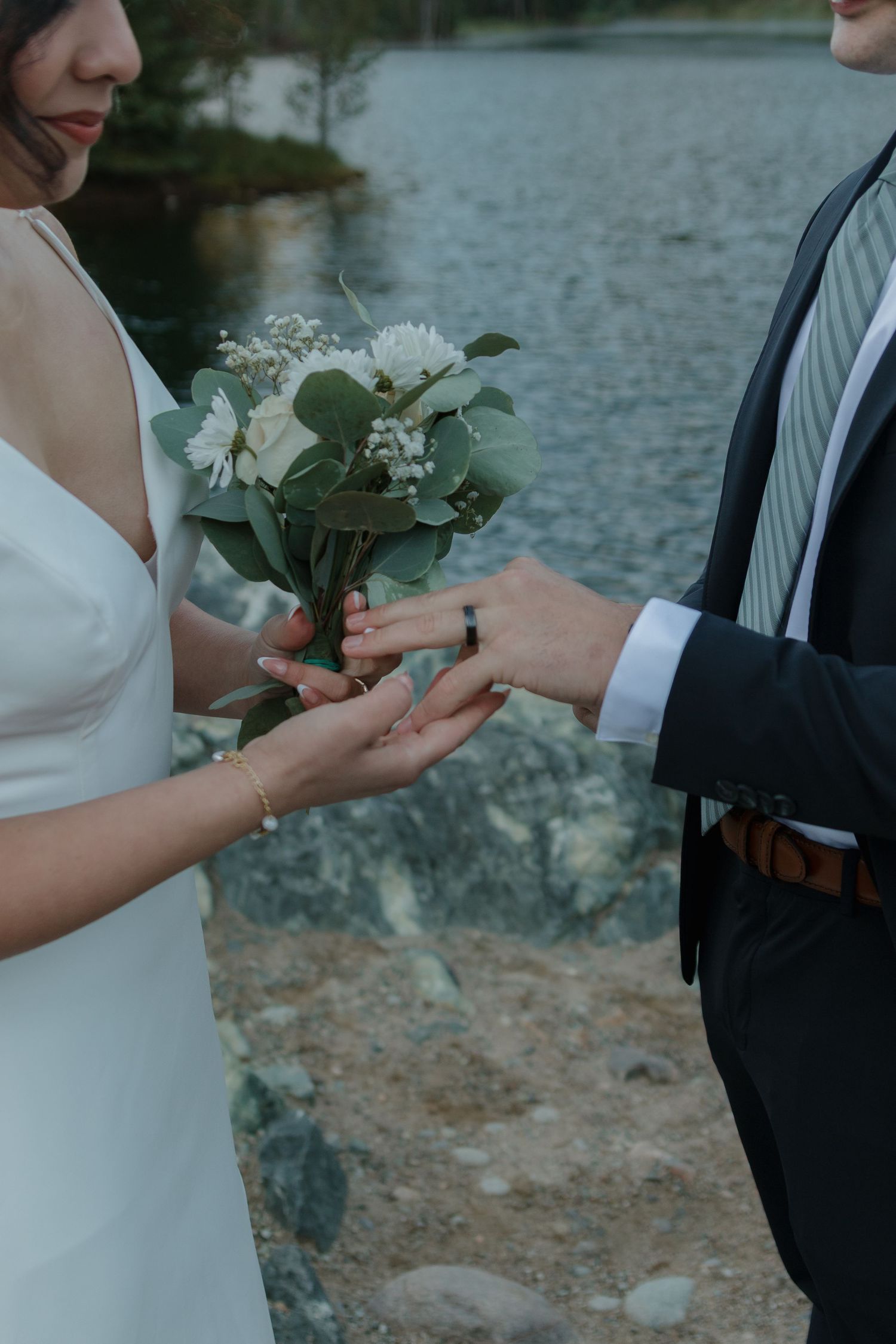 A couple exchanges white floral bouquets by a serene lake during their wedding ceremony.