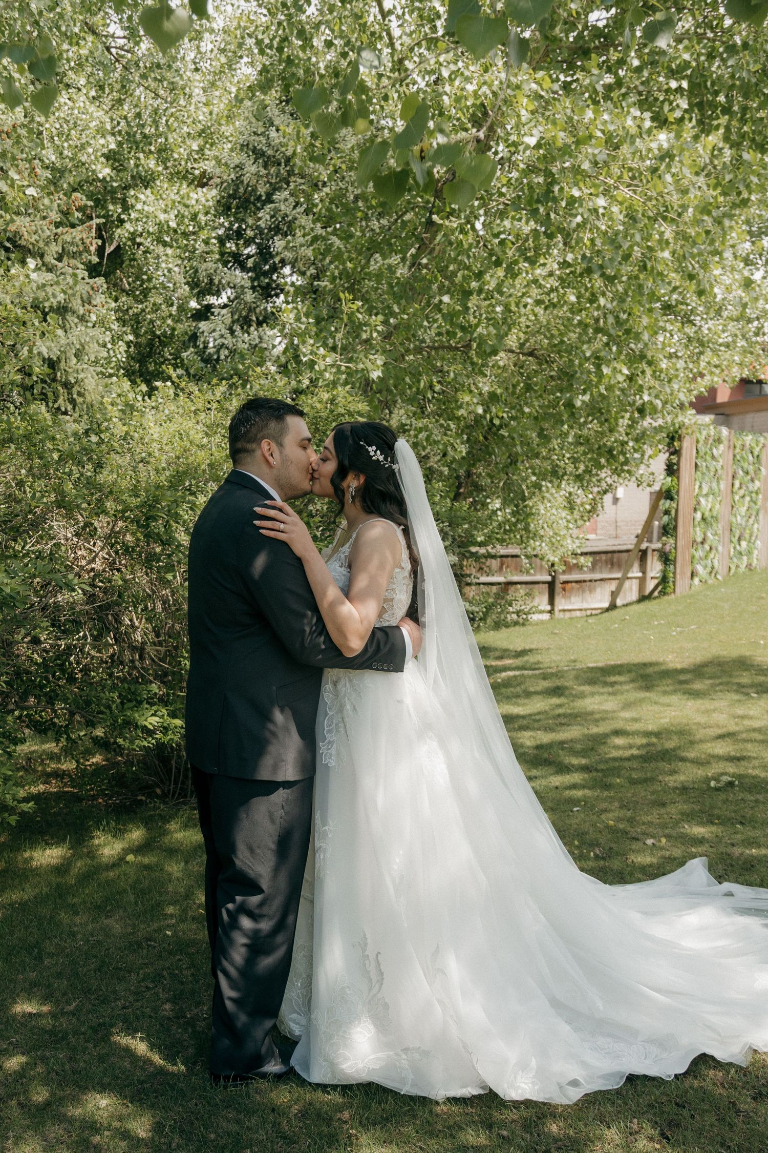 Bride and groom share a romantic kiss under lush green trees during outdoor wedding portrait session.