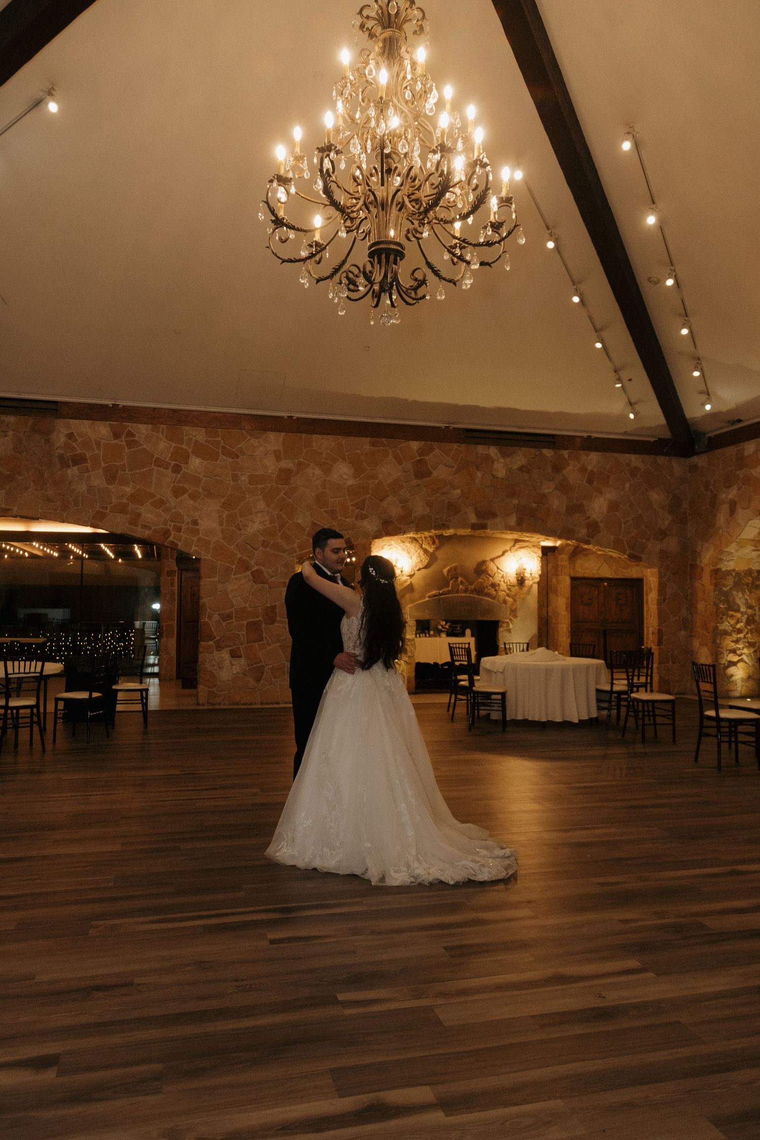 Bride and groom share romantic first dance under elegant chandelier in rustic stone wedding venue.