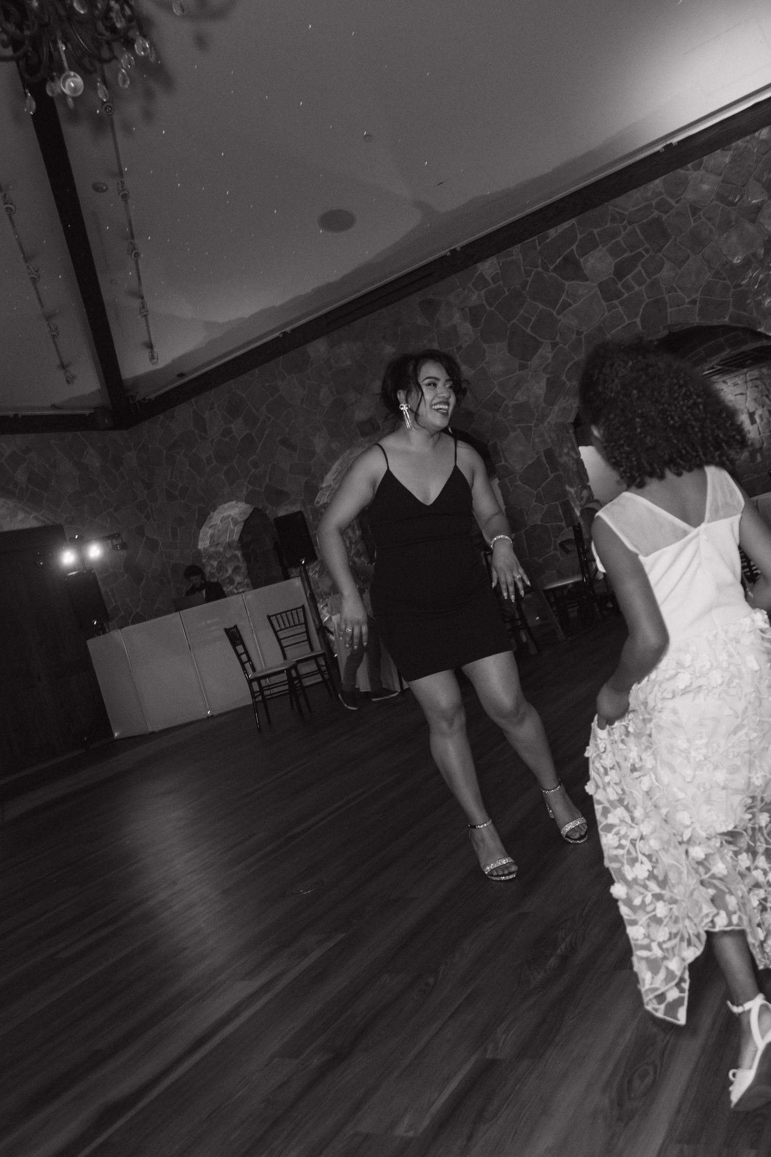 Two women dancing on a wooden dance floor in a stone-walled wedding reception venue, black and white photo.