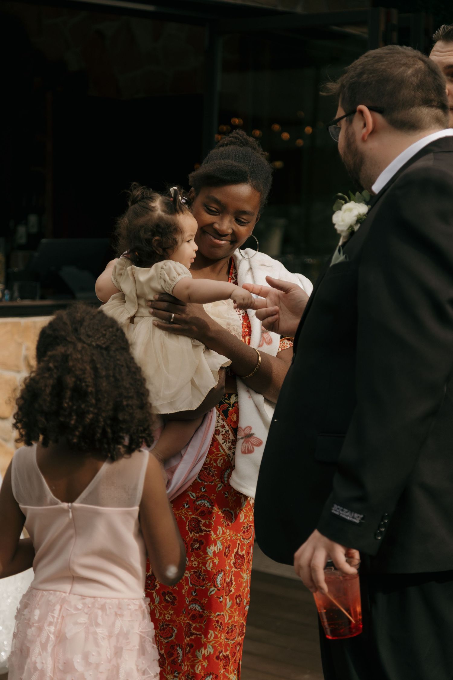Woman in floral dress holds baby while greeting suited guest at wedding reception.