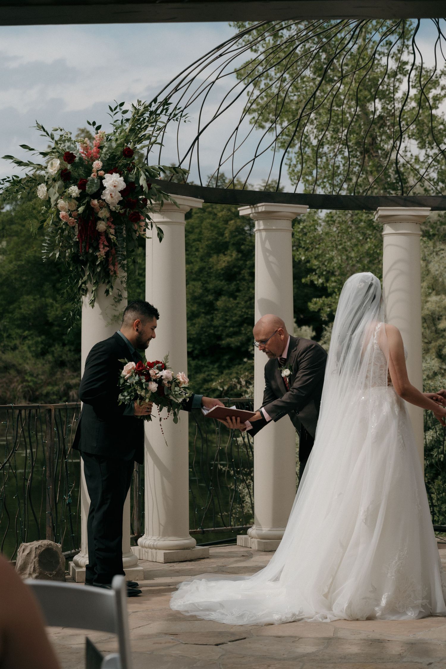 Bride and groom exchange vows under floral pergola with officiant at elegant outdoor wedding ceremony.