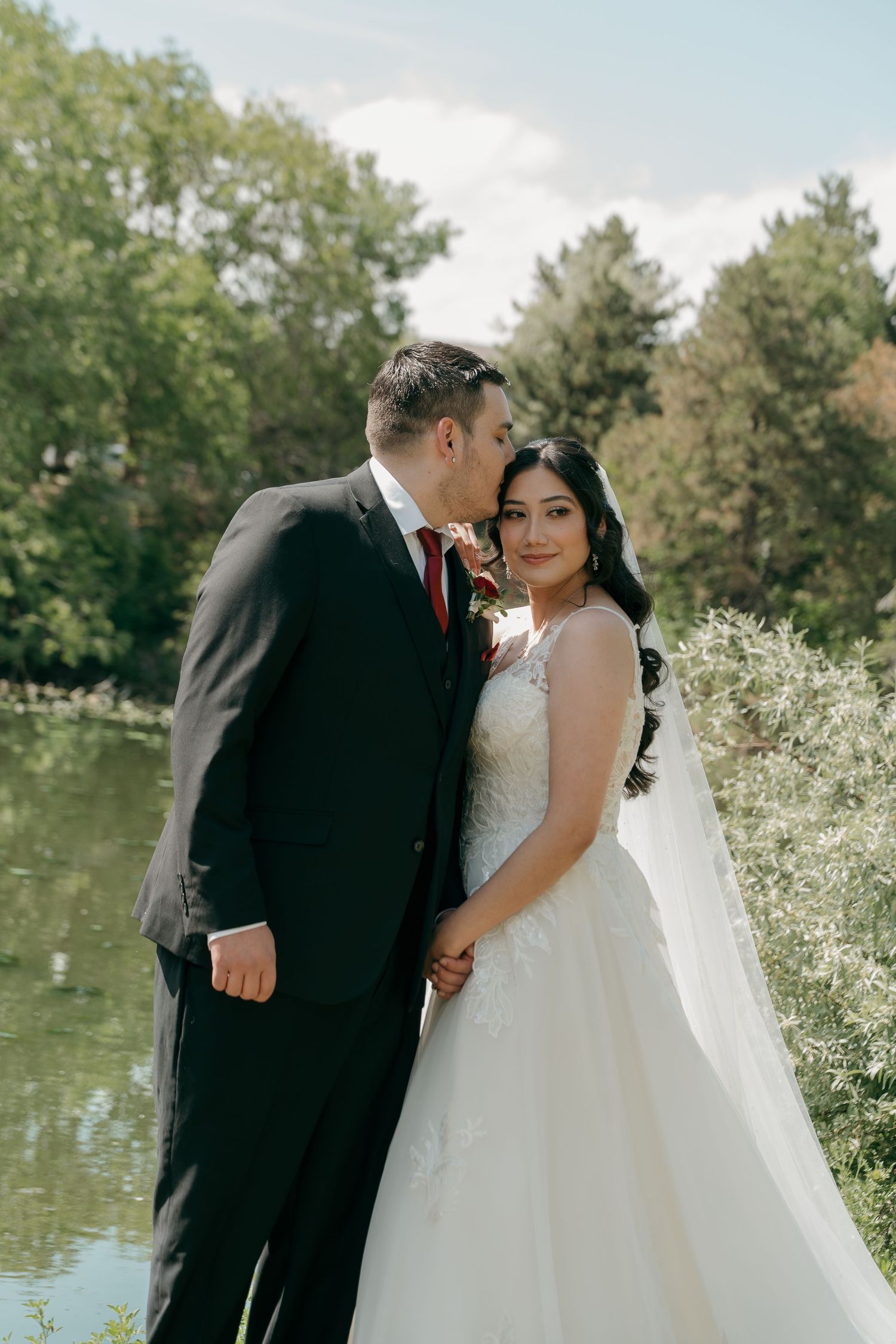 Bride and groom share a romantic moment by a serene pond surrounded by lush green trees on their wedding day.