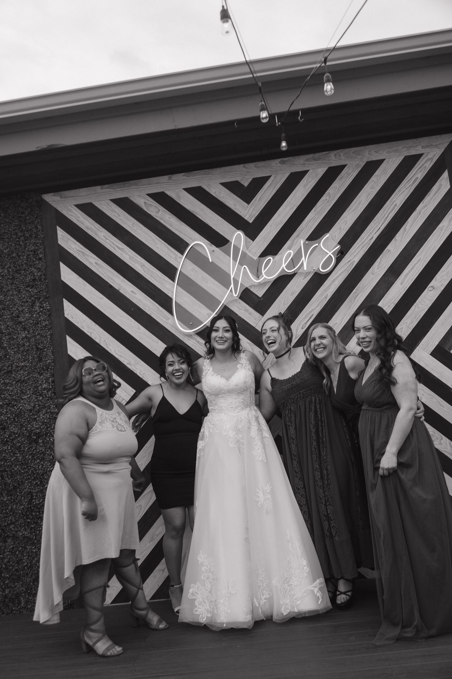 Bride poses with bridesmaids in front of a Cheers neon sign backdrop at wedding reception.