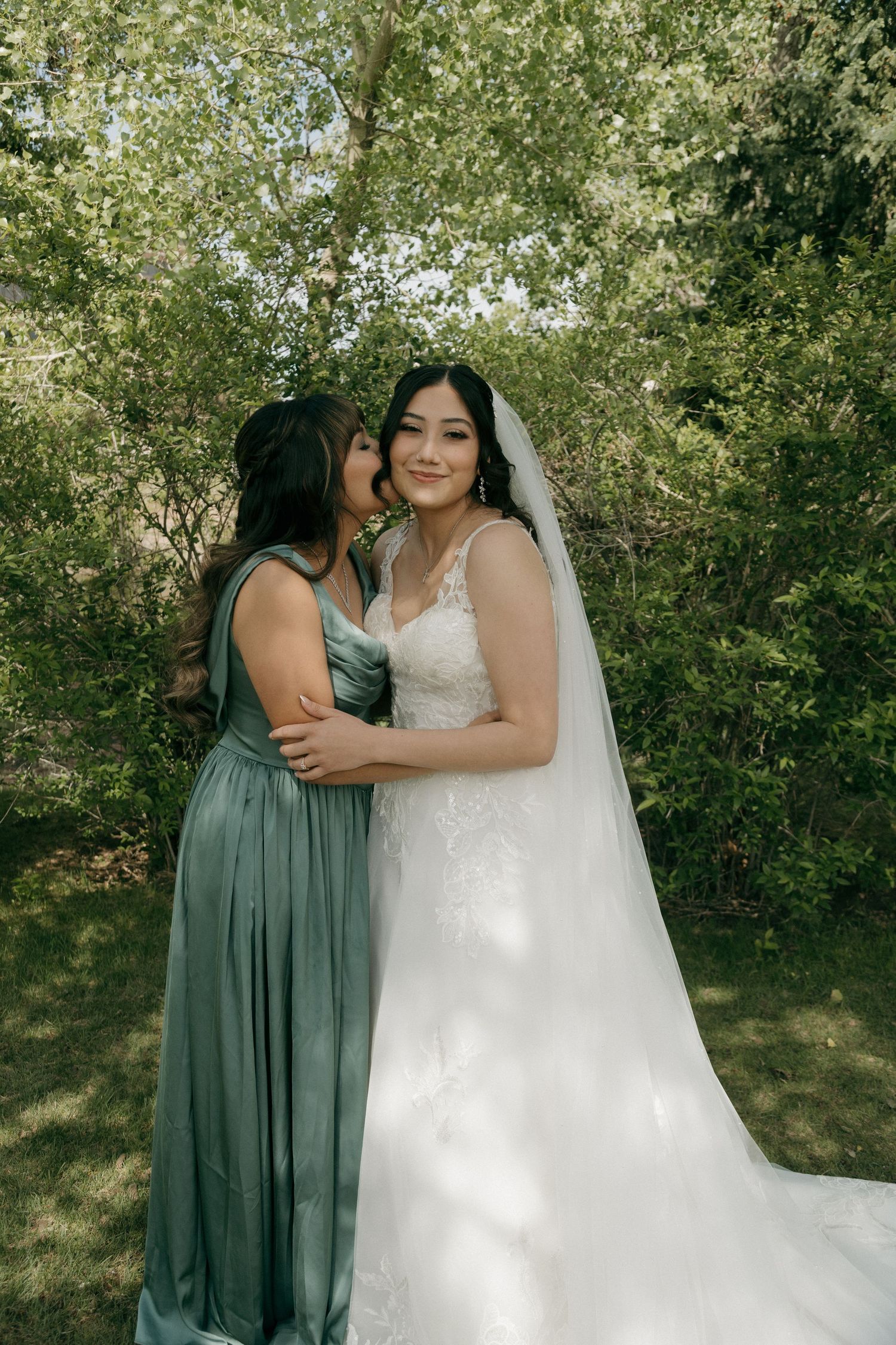Bride in lace wedding gown and veil embraces bridesmaid in sage green dress outdoors among green trees.