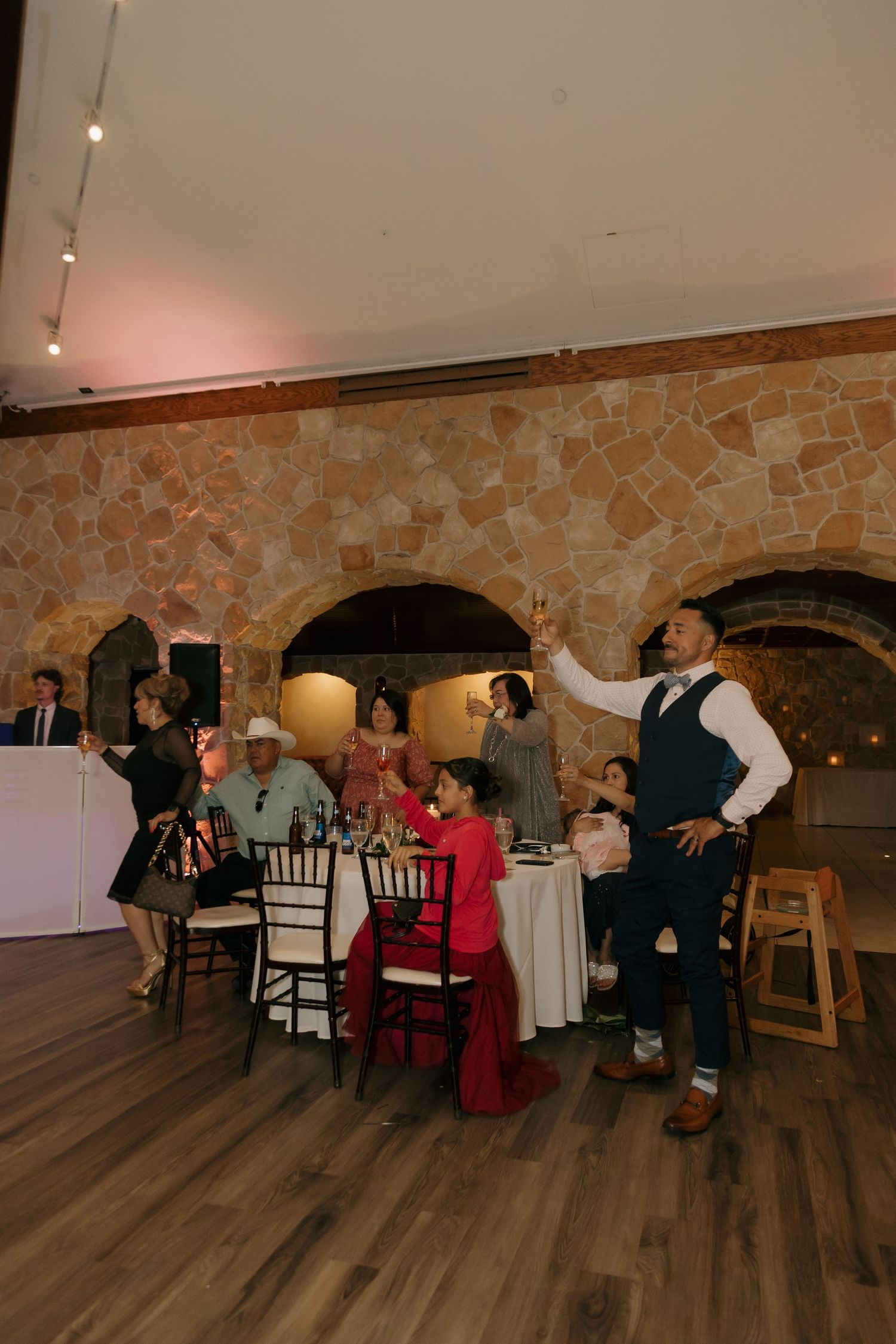 Wedding guest raises a champagne toast in a rustic stone-walled reception hall with warm lighting.