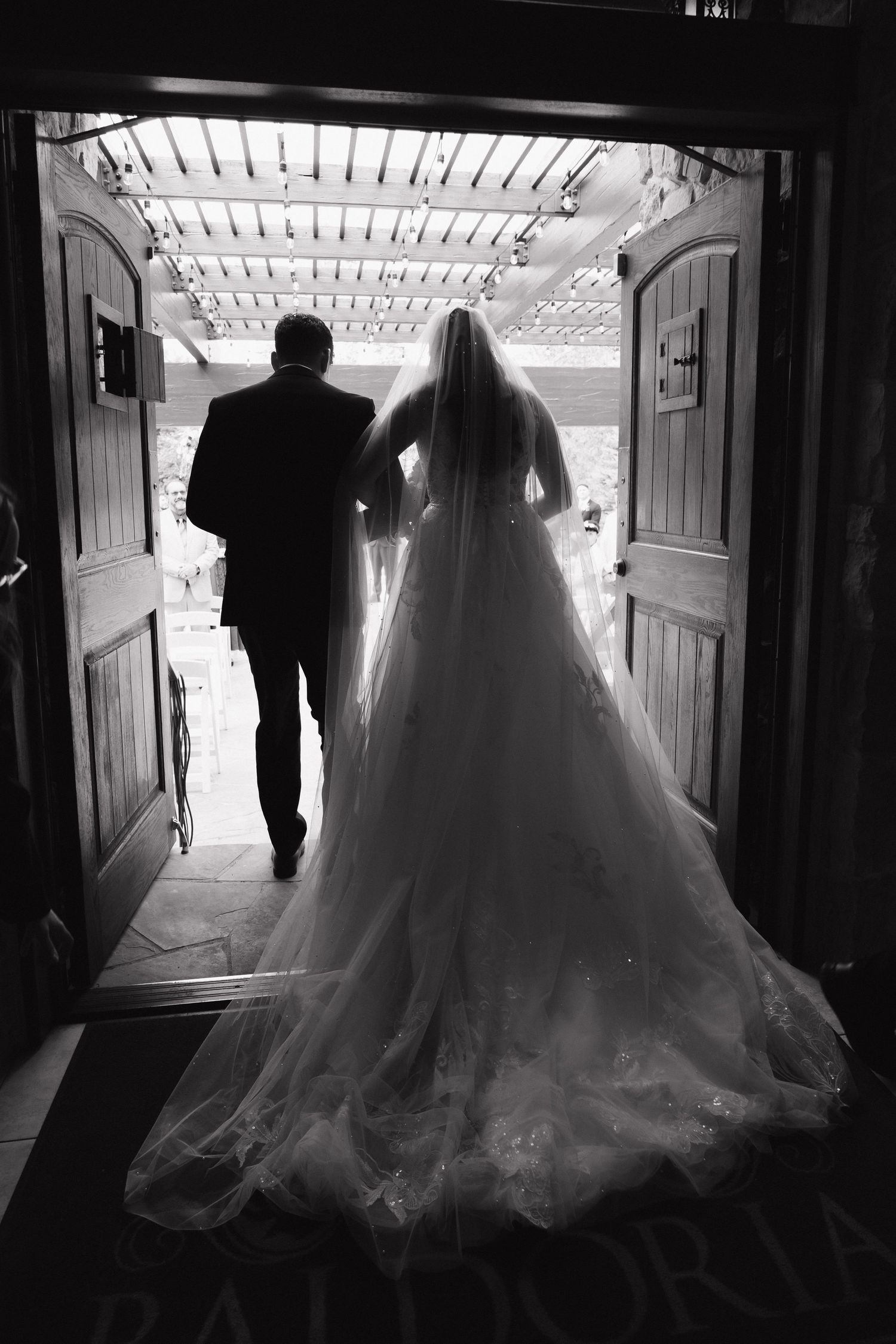 Black and white silhouette of bride in ballgown with escort walking through venue doors.