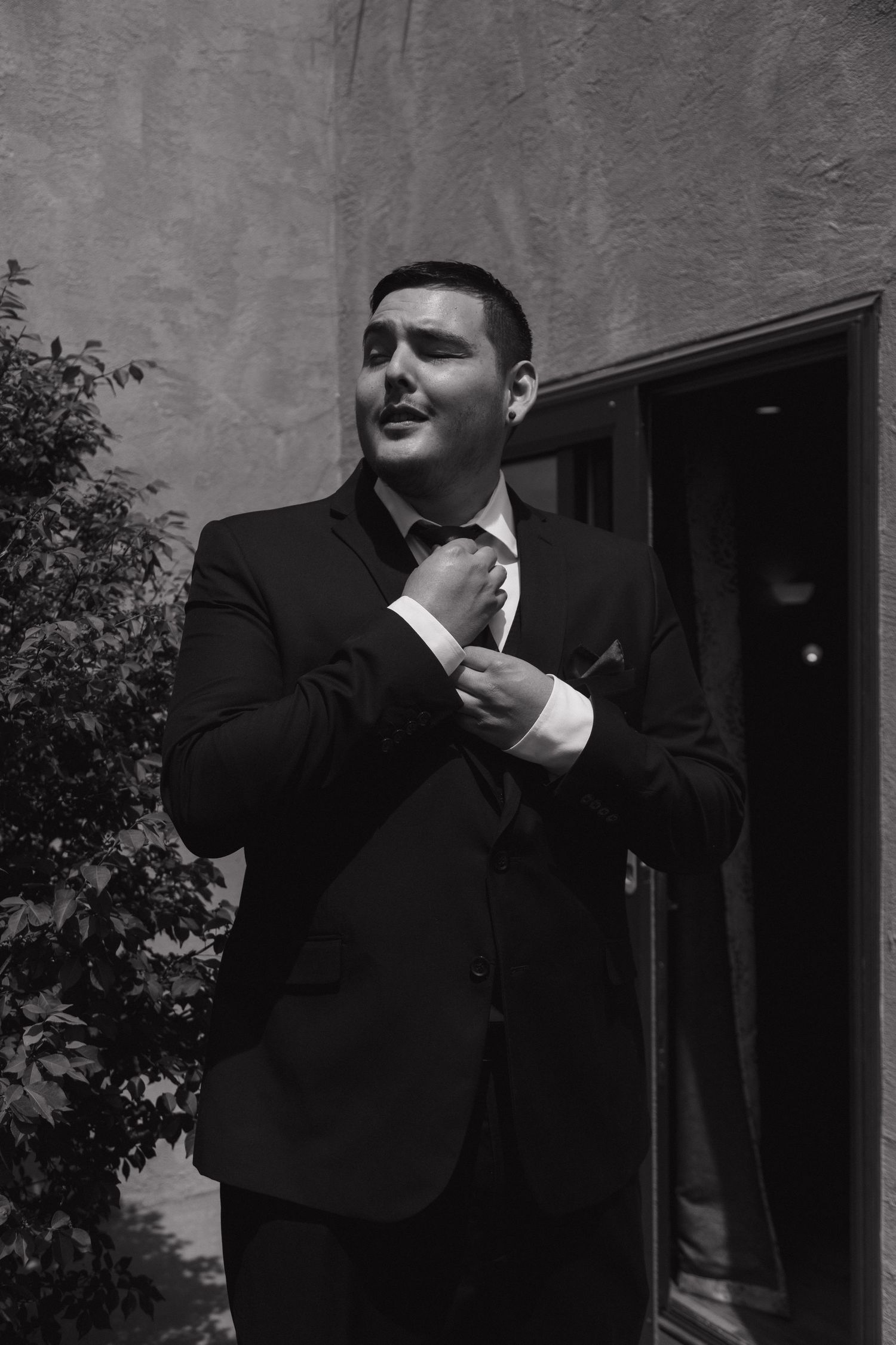 A groom in a black suit adjusts his tie outdoors in a black and white wedding photo.