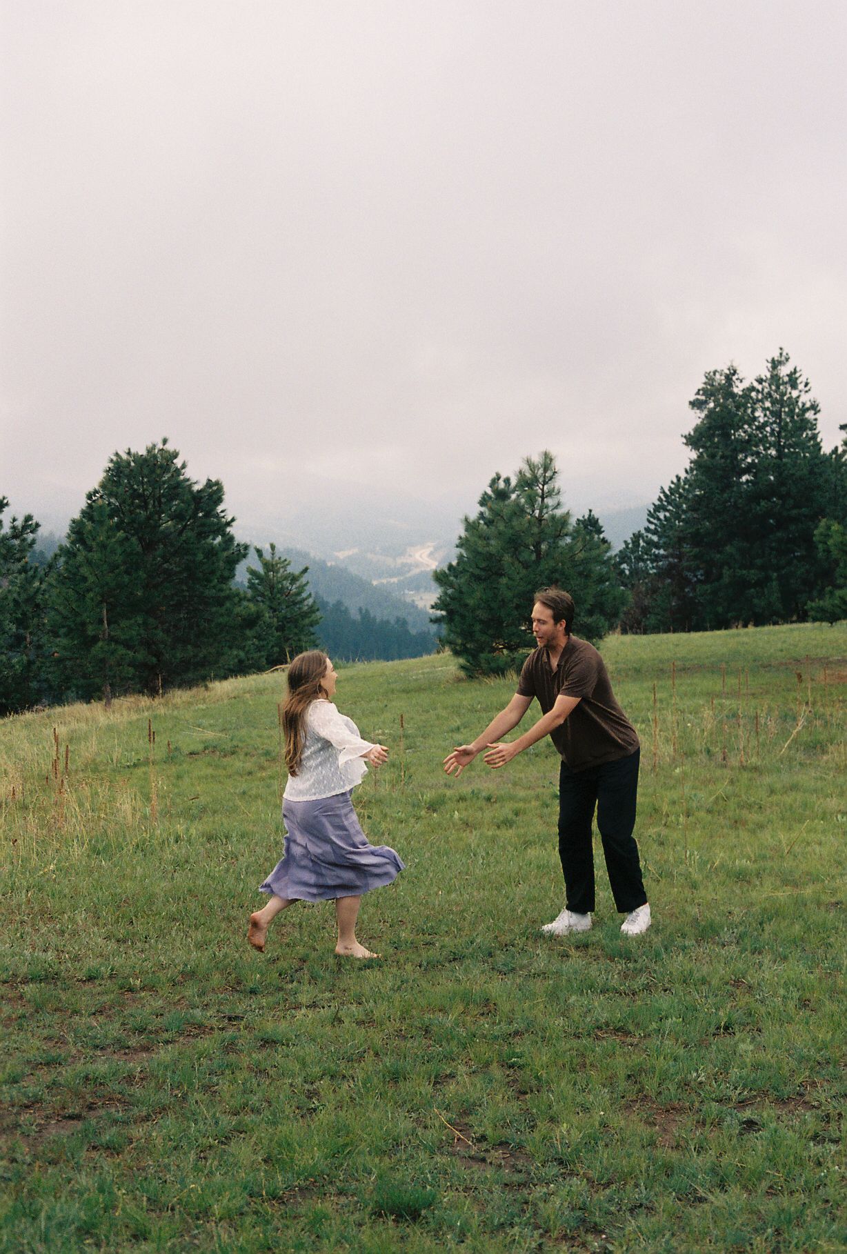Playful couple running toward each other in a lush green mountain meadow with pine trees.