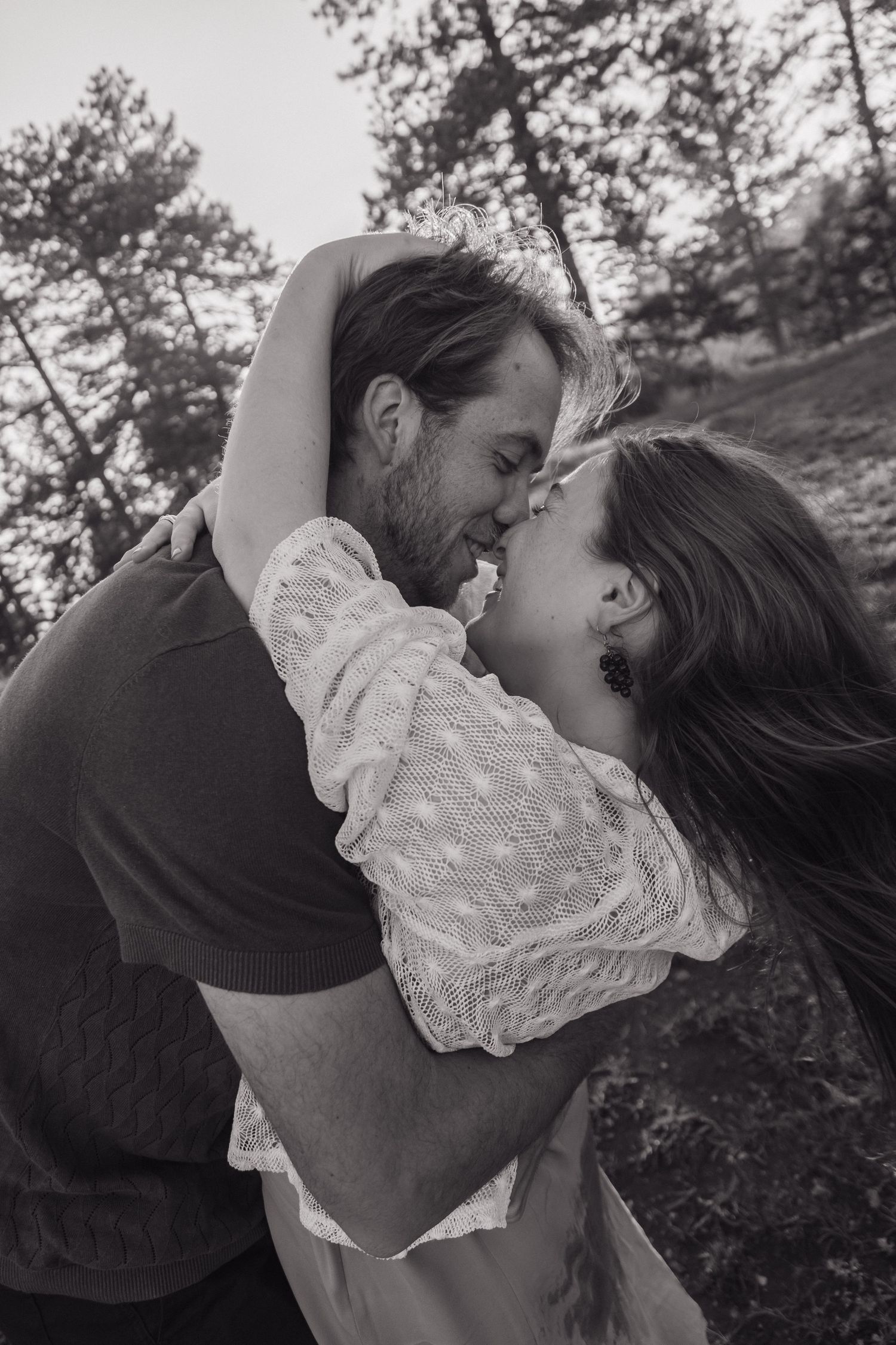 Black and white photo of a couple sharing a passionate kiss outdoors among pine trees.