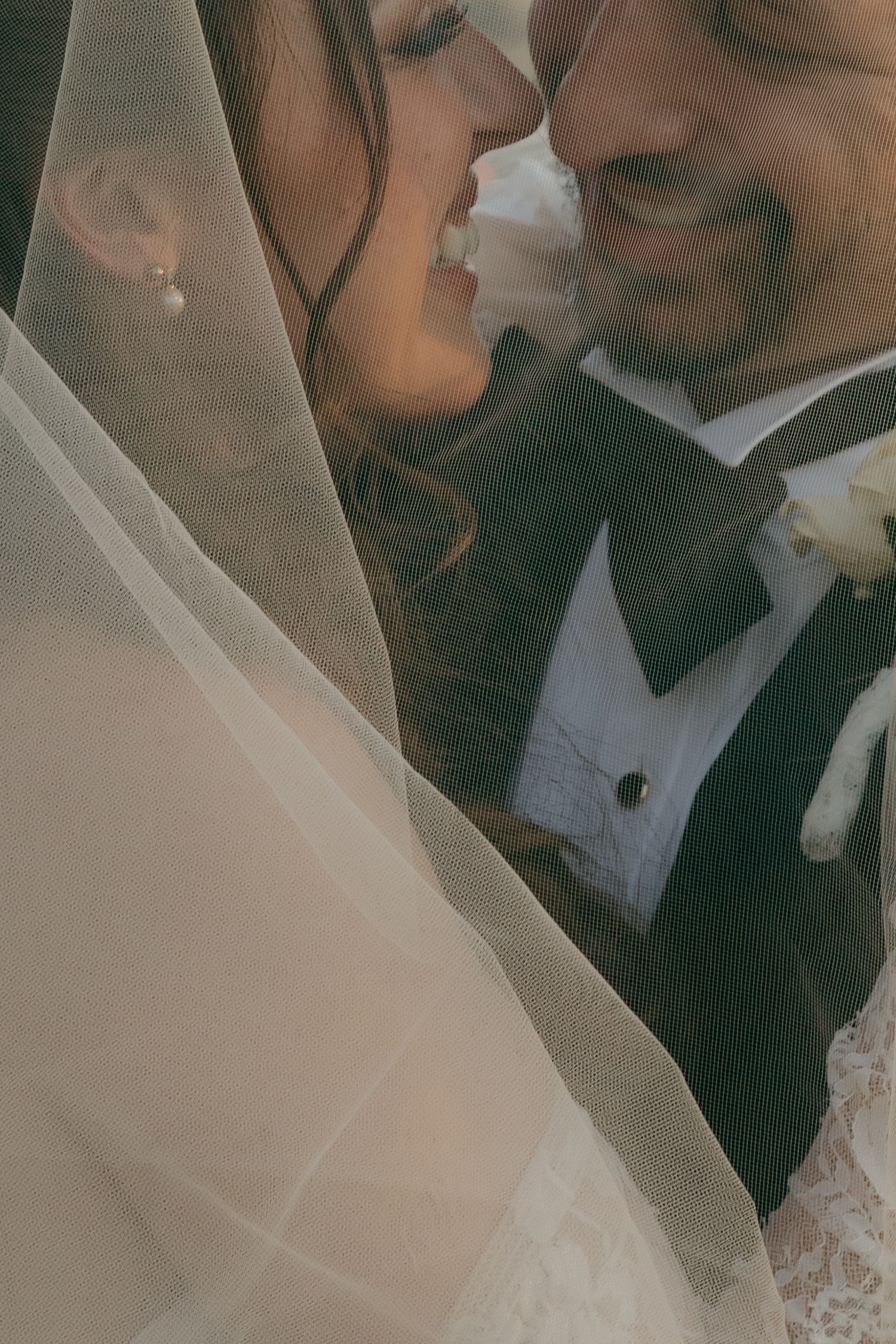 Bride and groom share an intimate moment under a flowing wedding veil, smiling closely together.
