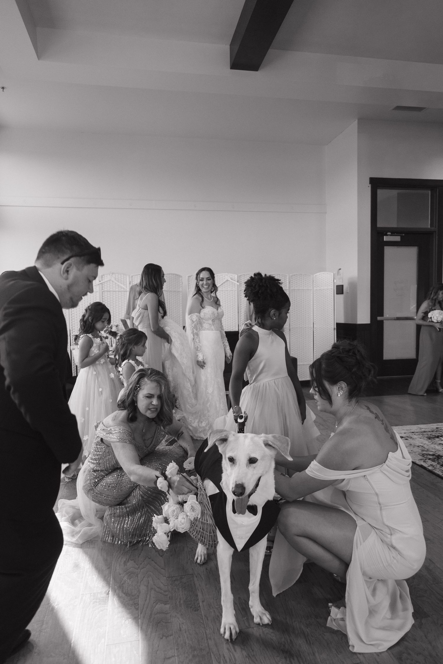 Wedding party gathers around dog wearing tuxedo bow tie in black and white pre-ceremony photo.