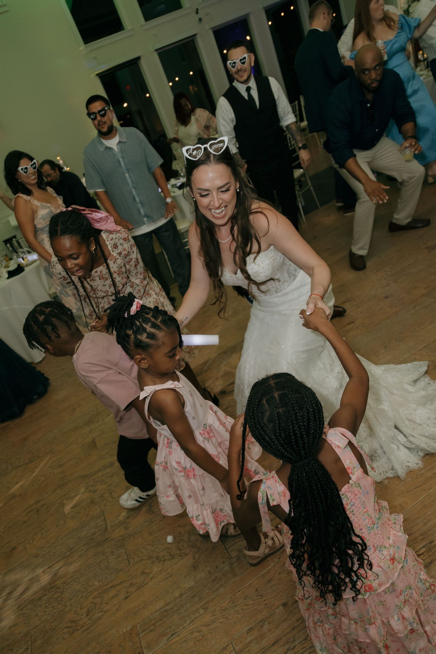 Bride in lace gown with heart sunglasses dances joyfully with young flower girls at wedding reception.