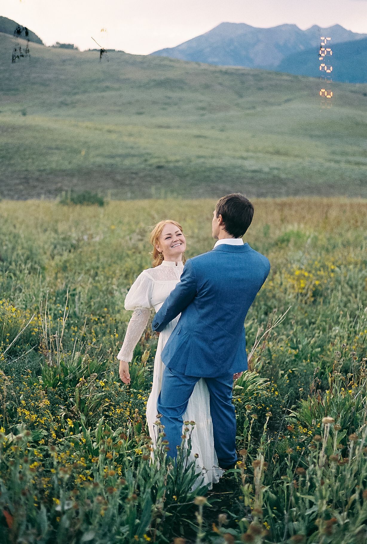 Smiling bride in white gown dances with groom in blue suit in wildflower meadow.