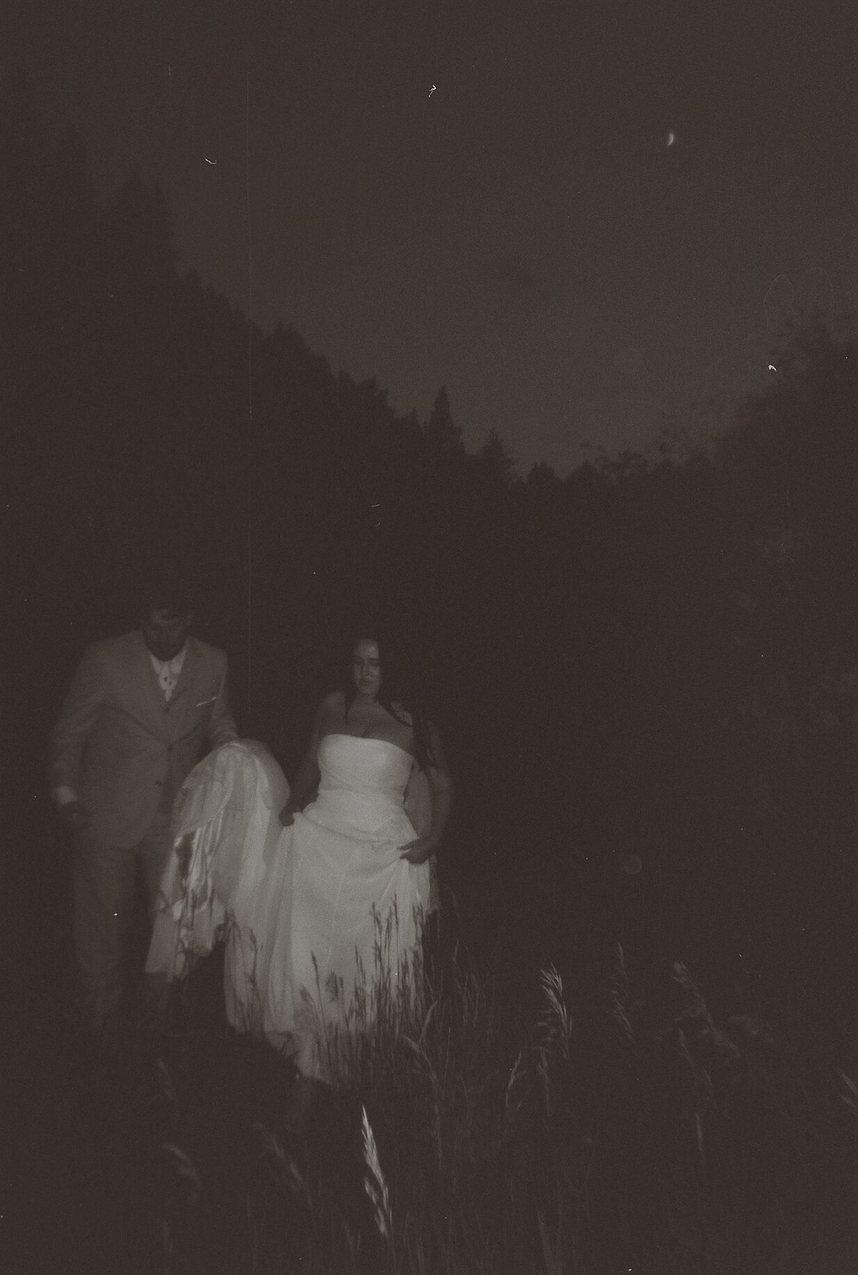 Romantic dark film photo of bride and groom walking under starry night sky in forest.