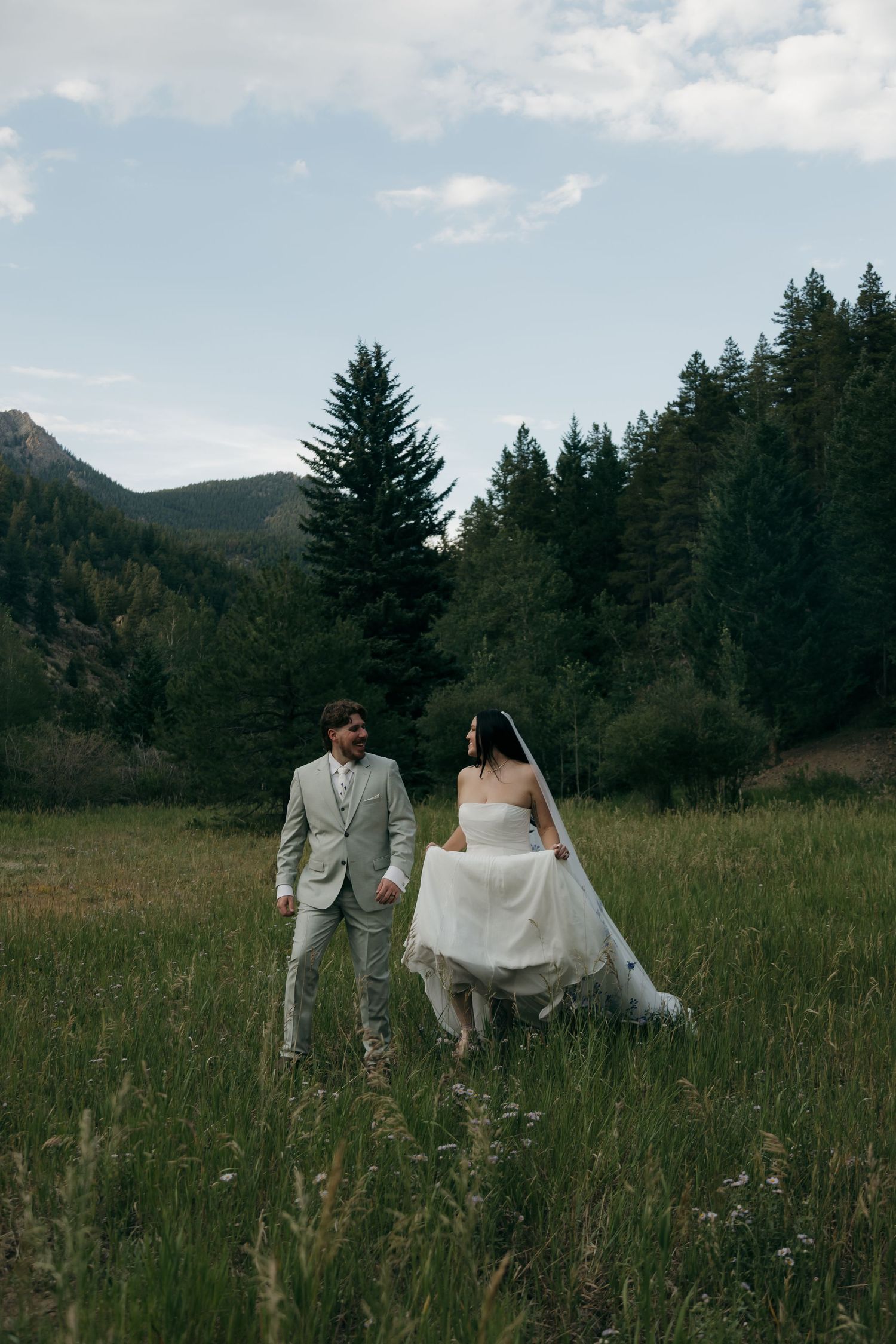 Bride in a flowing white gown and groom in a light gray suit walk through a scenic mountain meadow surrounded by evergreens.