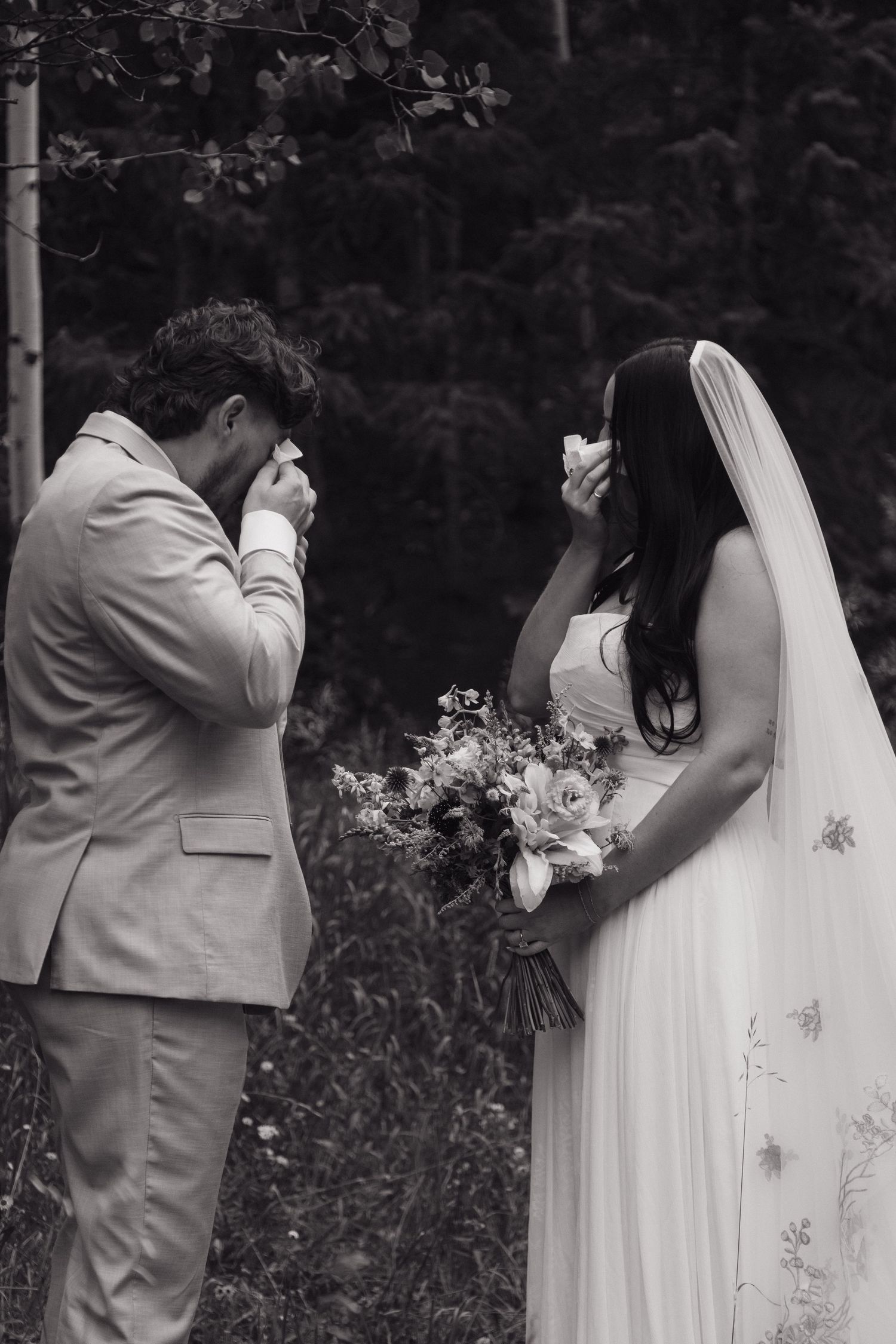 Black and white photo of bride and groom wiping tears during outdoor forest wedding ceremony with bouquet.