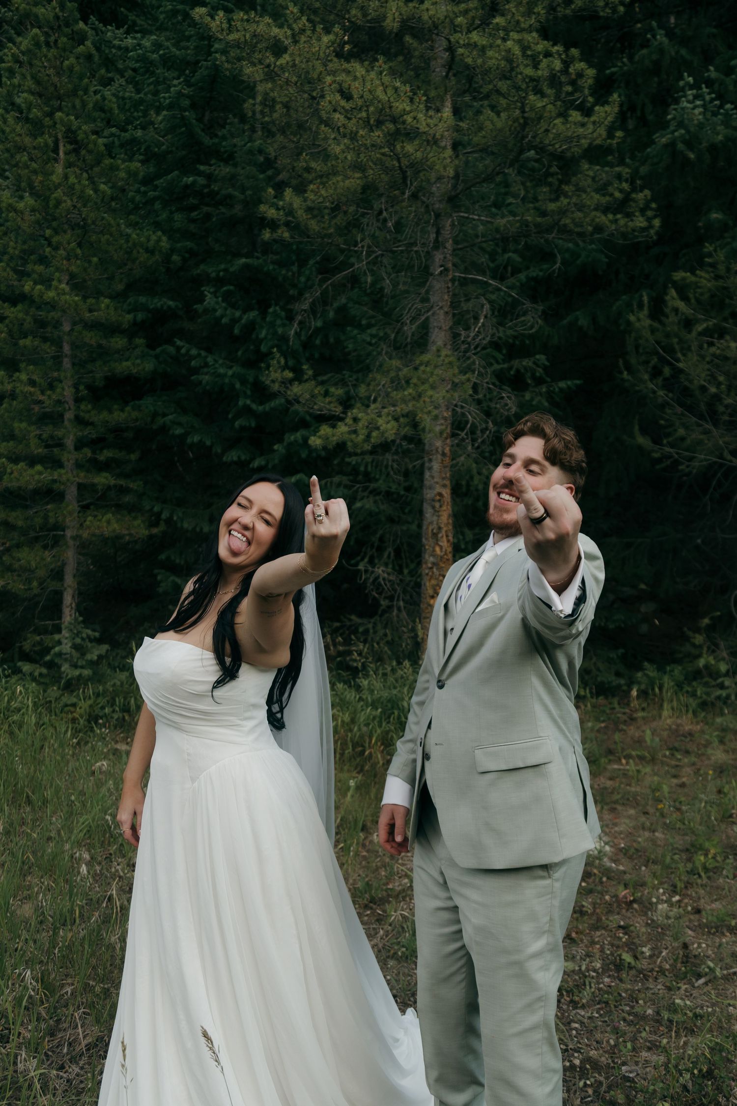 Joyful bride and groom in sage suit laugh and point in a lush forest meadow on their wedding day.