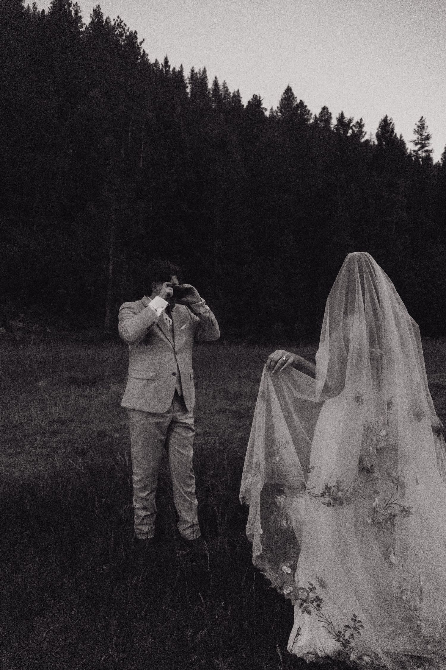 Black and white photo of groom photographing veiled bride in forest meadow at twilight.