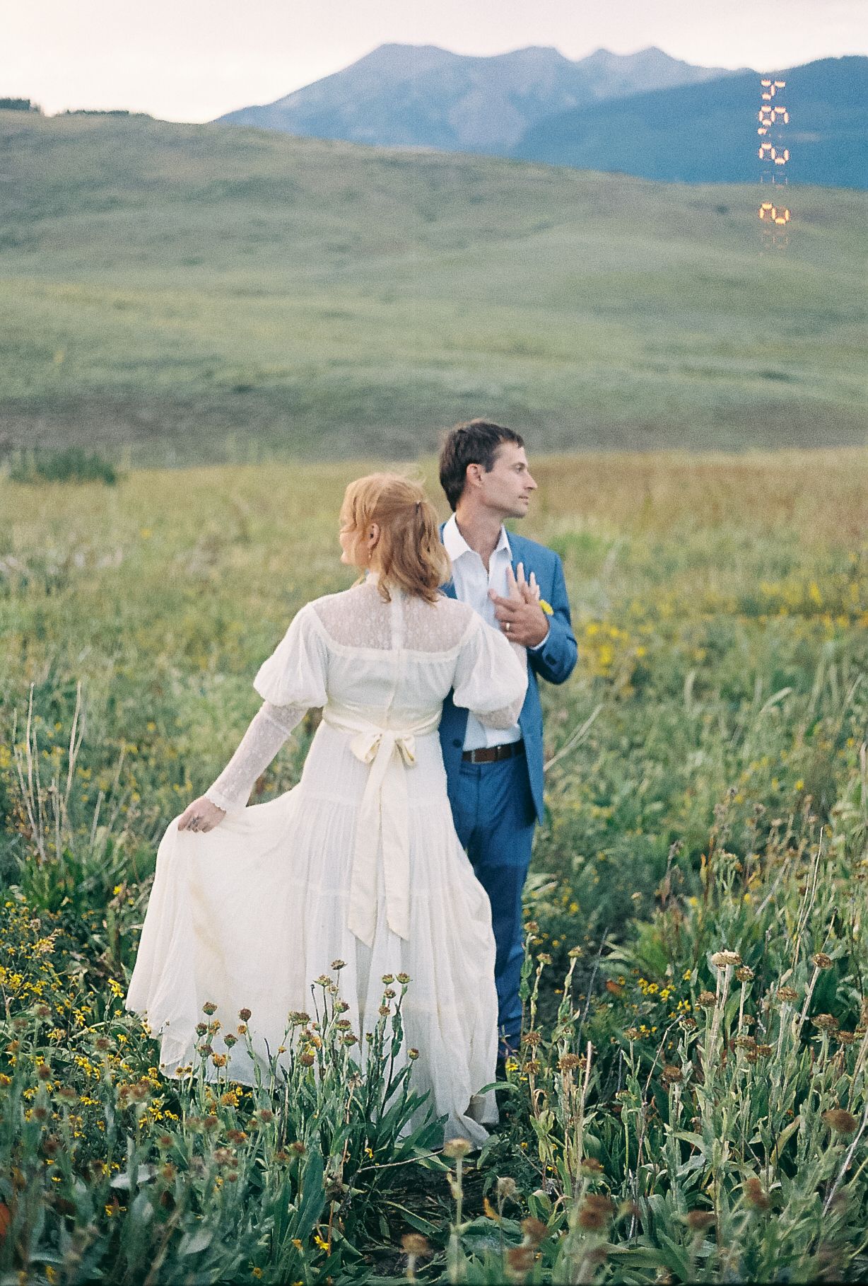 Bride in white dress with bow and groom in blue suit walk through mountain wildflower meadow at dusk.