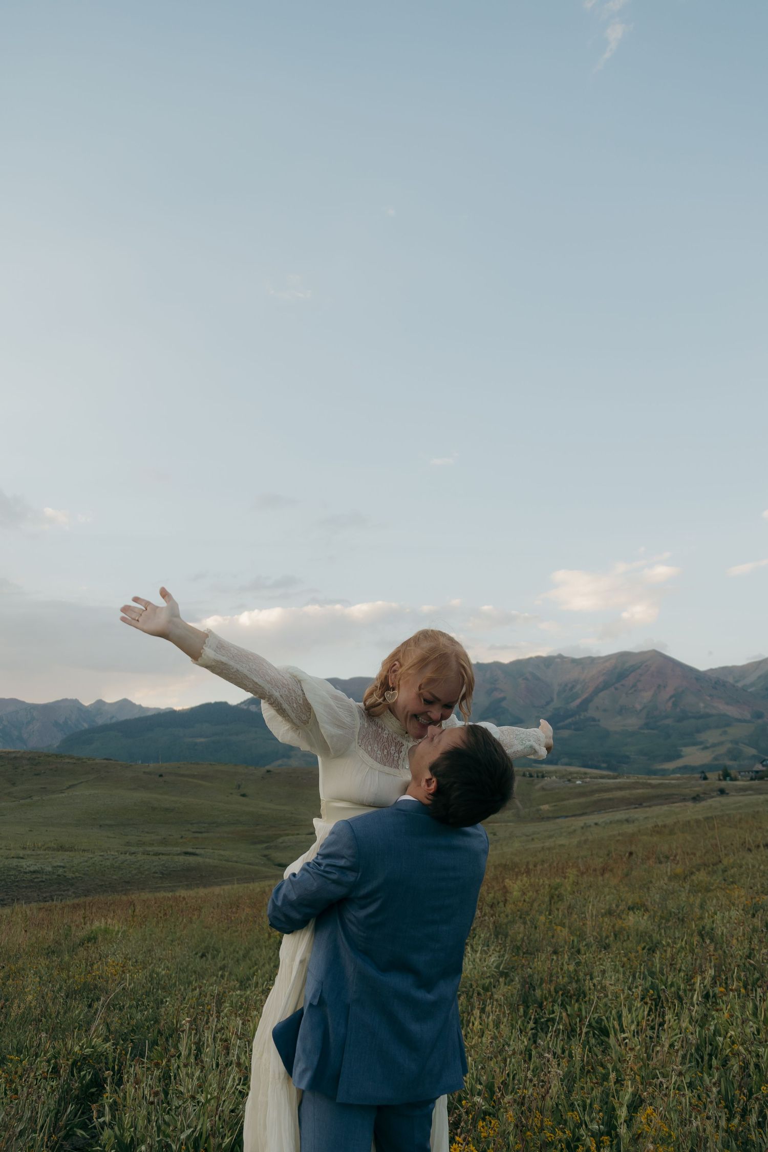 Groom lifts bride in white dress against scenic mountain meadow backdrop at golden hour wedding.
