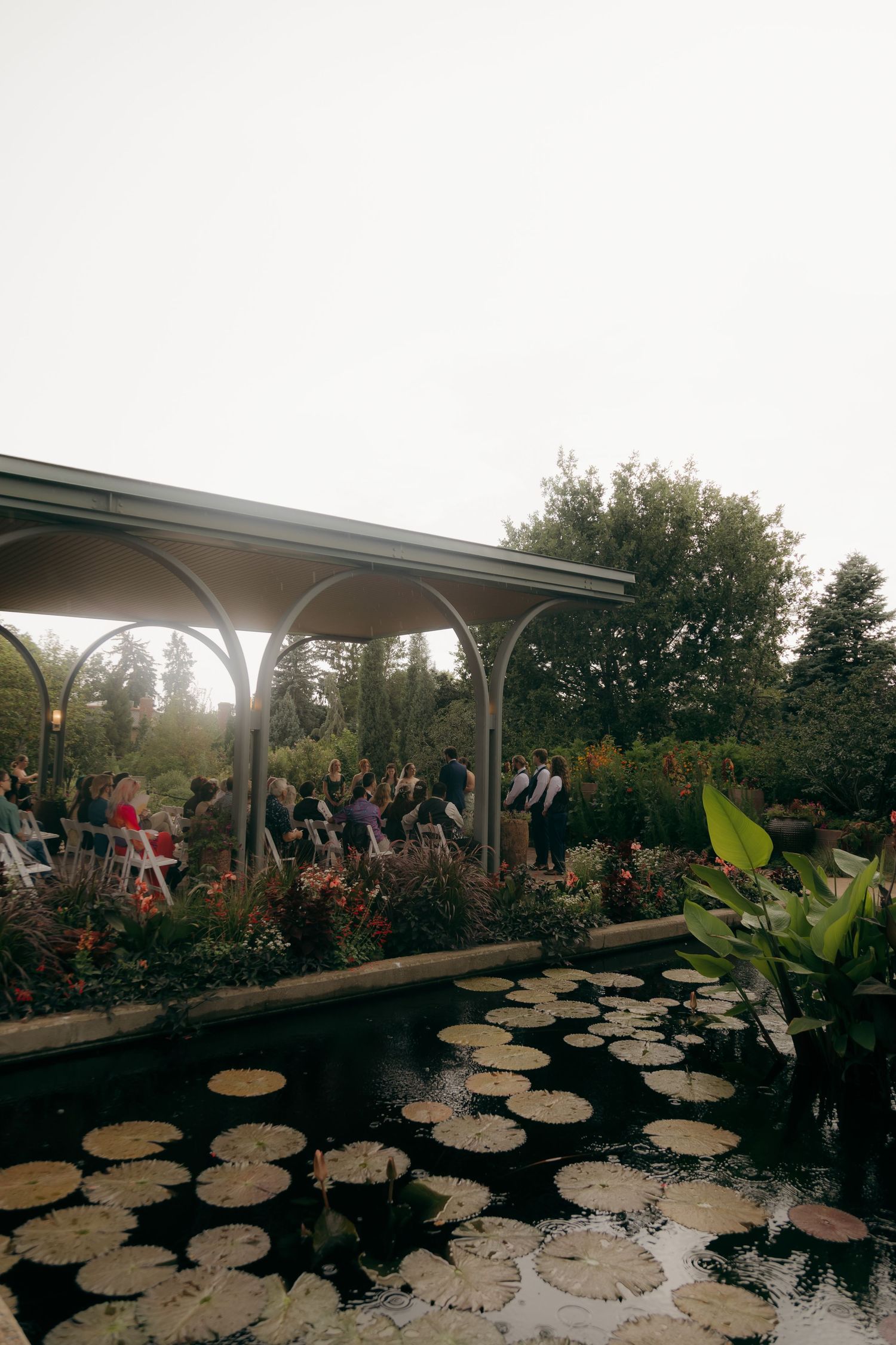 Outdoor wedding ceremony under a pergola beside a tranquil pond with lily pads, surrounded by lush garden greenery.