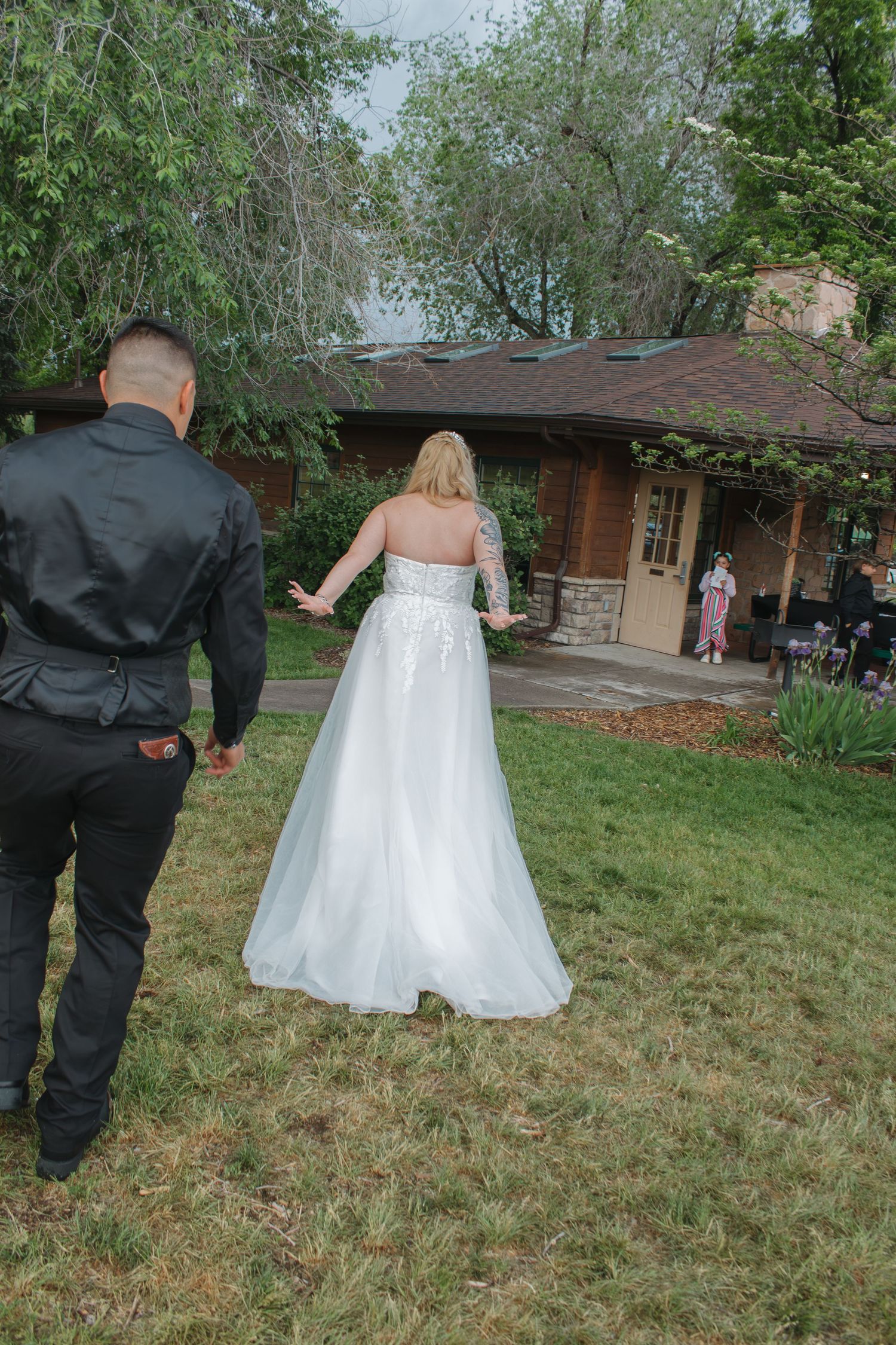 Bride in white gown and person in black suit walking through green lawn near brick house.