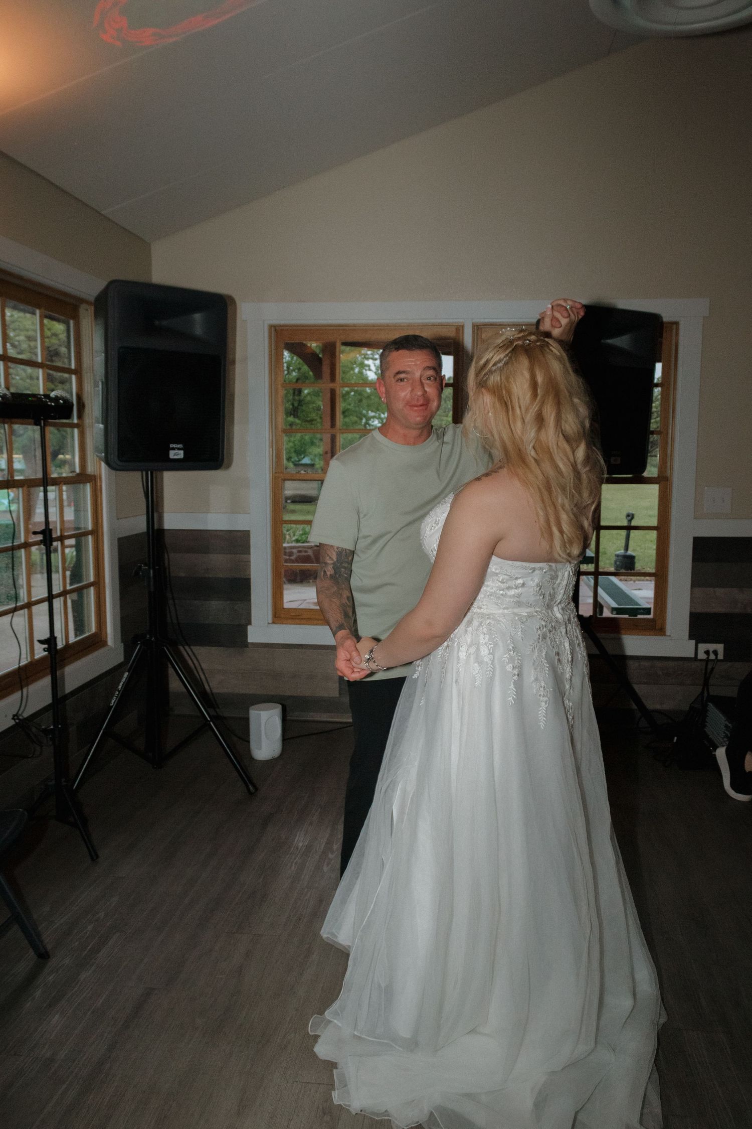 Couple dancing together in a bright room with windows and wooden flooring during what appears to be a reception.