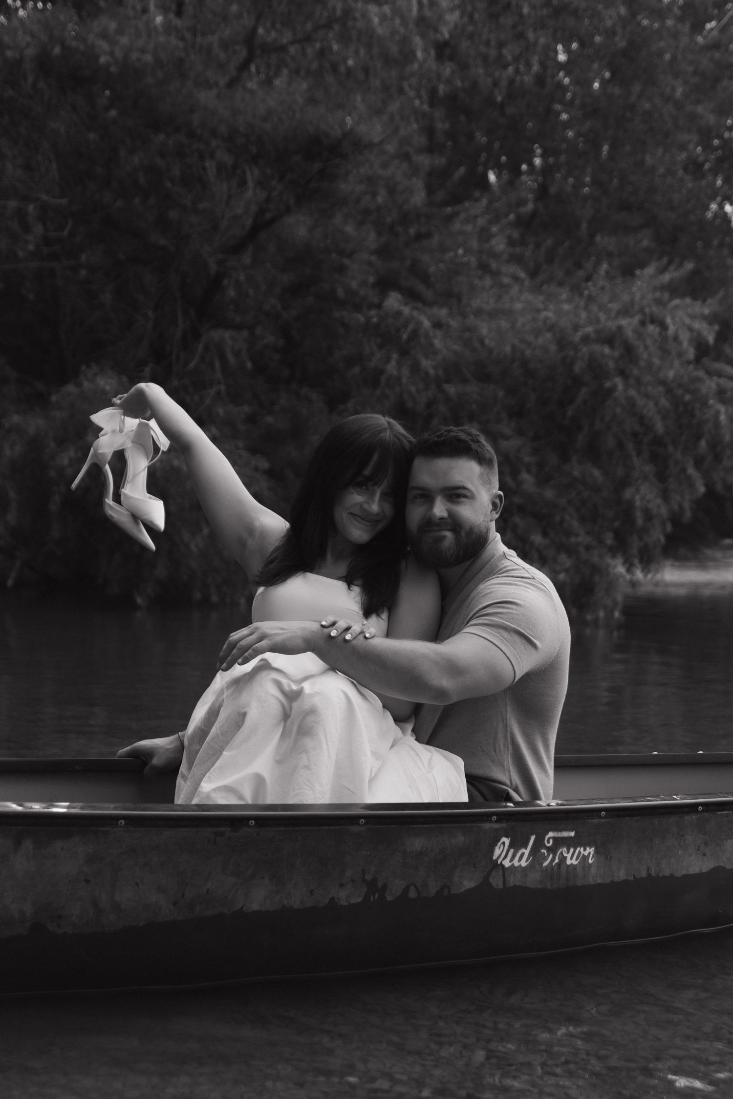 A romantic couple sits close together in a canoe on a tranquil lake surrounded by trees.